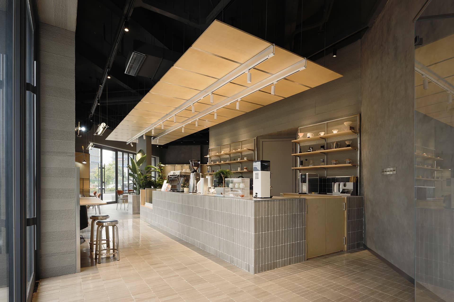 Modern minimalist cafe interior featuring a long grey concrete tile service counter, wooden canopy lighting fixture, metal bar stools, and open shelving against concrete walls.