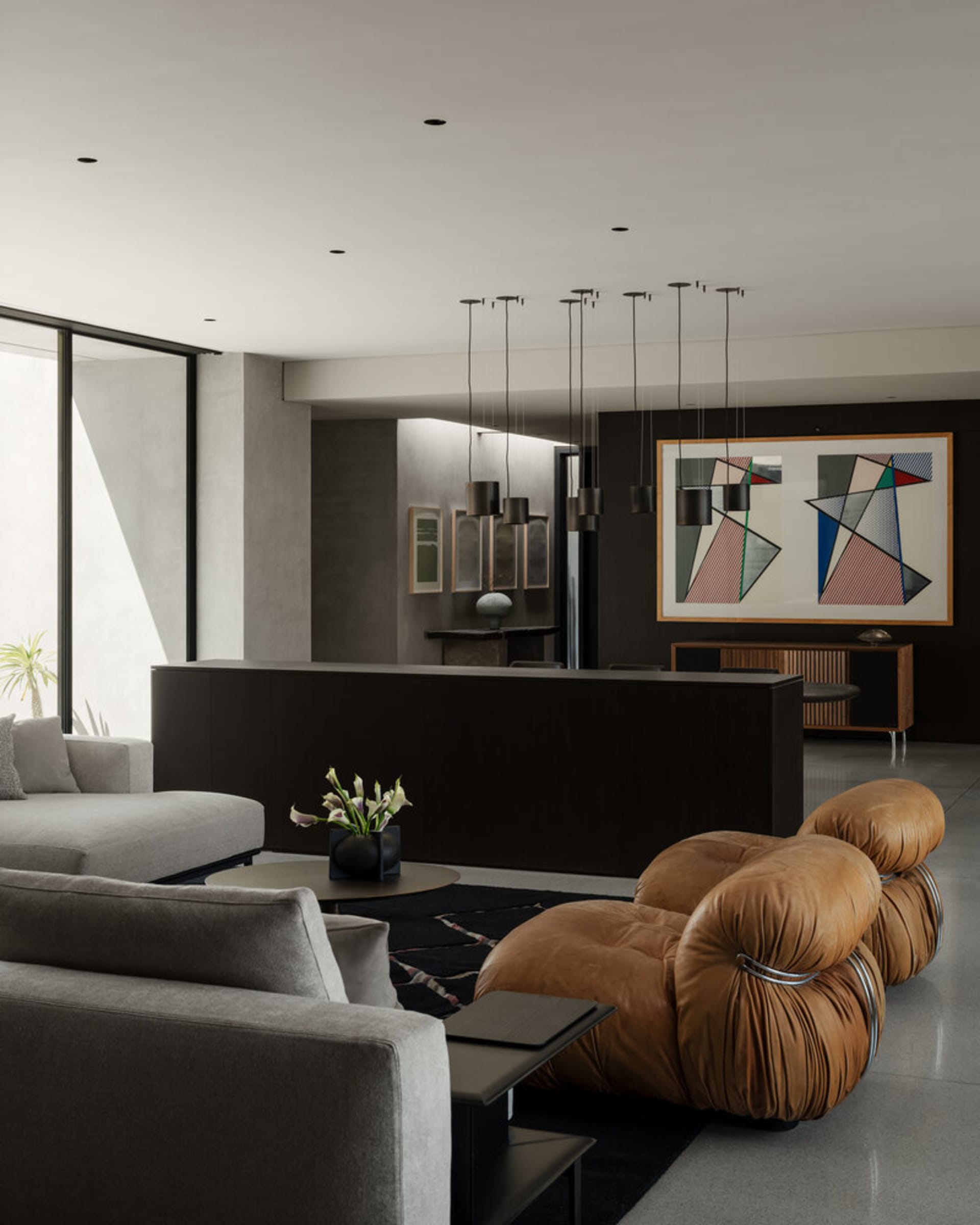 Modern living room featuring a brown leather bubble sofa, grey sectional, dark wood console, large abstract geometric painting, and multiple pendant lights.