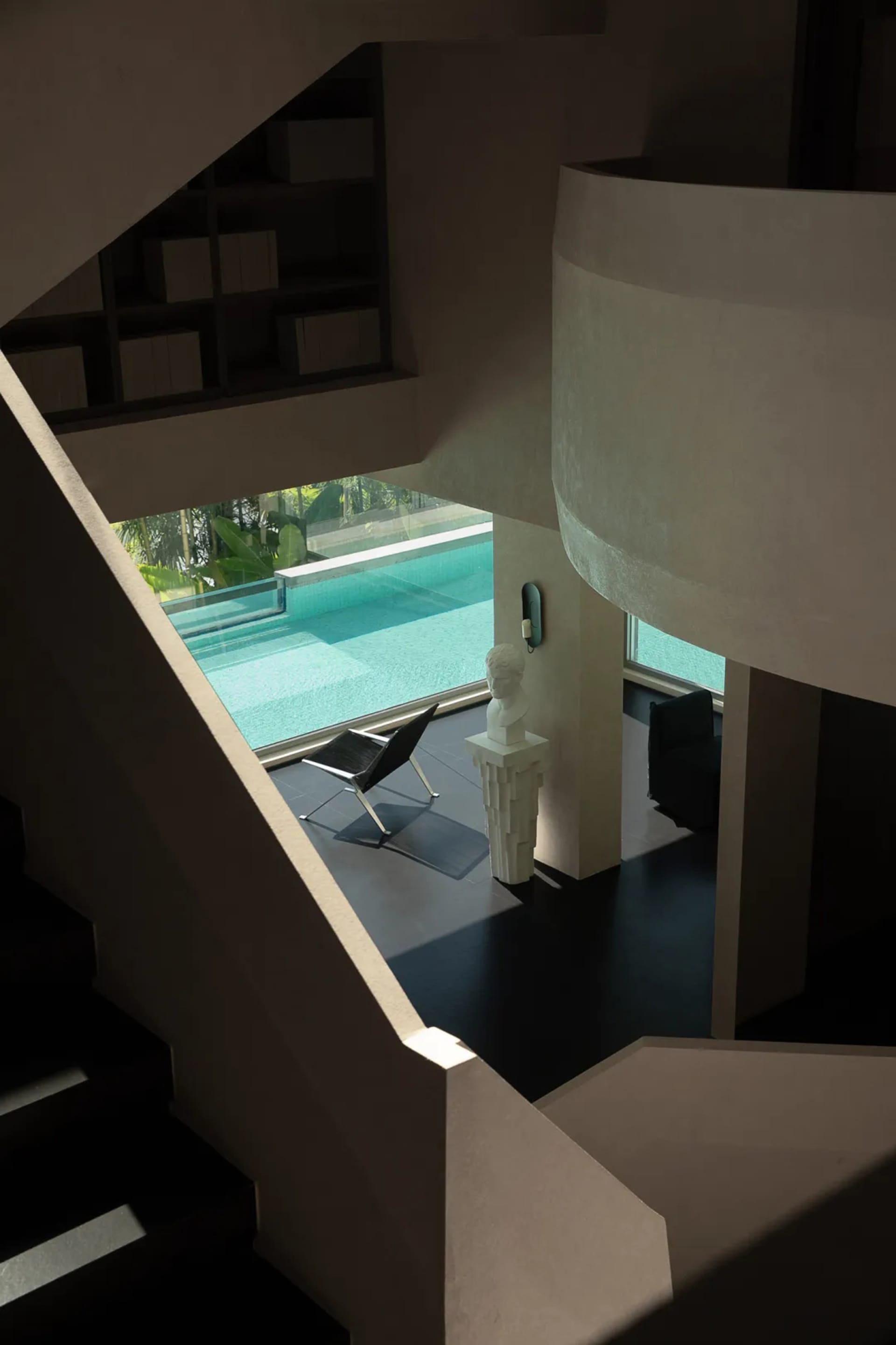 View from above of a modern curved concrete staircase overlooking a contemporary living area with a white classical bust on a pedestal, black leather chair, and large glass windows revealing an infinity pool surrounded by tropical greenery.