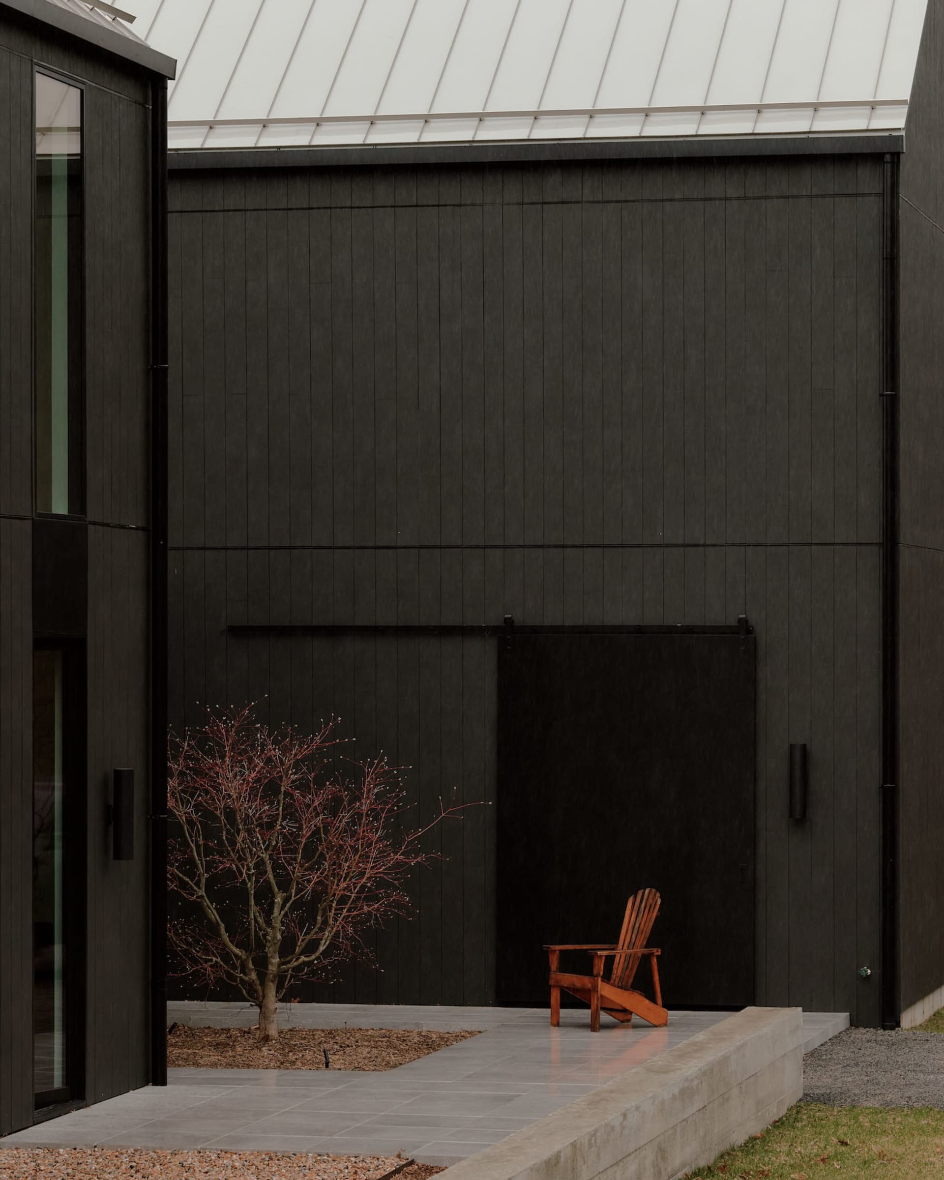 Modern black charred wood house exterior with concrete patio, small red-leaf tree, and orange Adirondack chair in a minimalist courtyard.