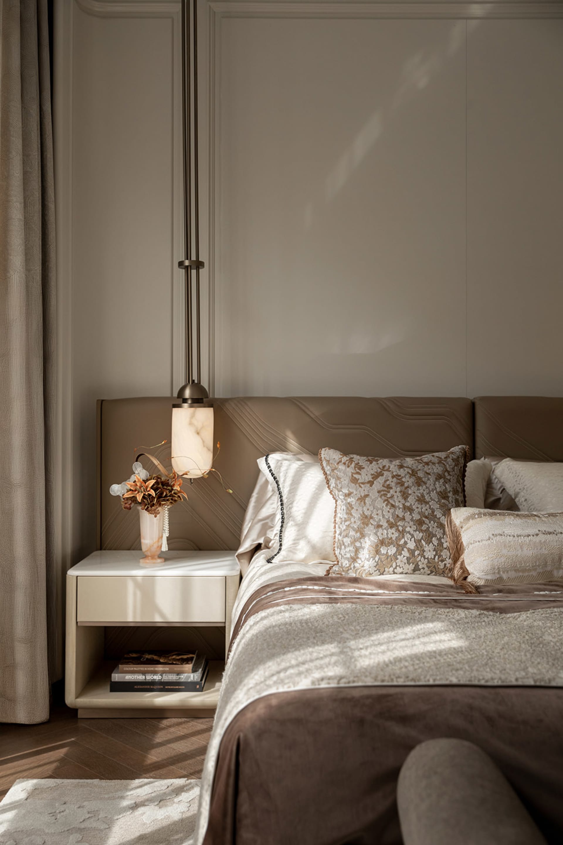 Modern luxury bedroom featuring a taupe leather upholstered headboard, marble pendant lamp, neutral bedding with floral accent pillows, and a white marble-top nightstand with dried floral arrangement.