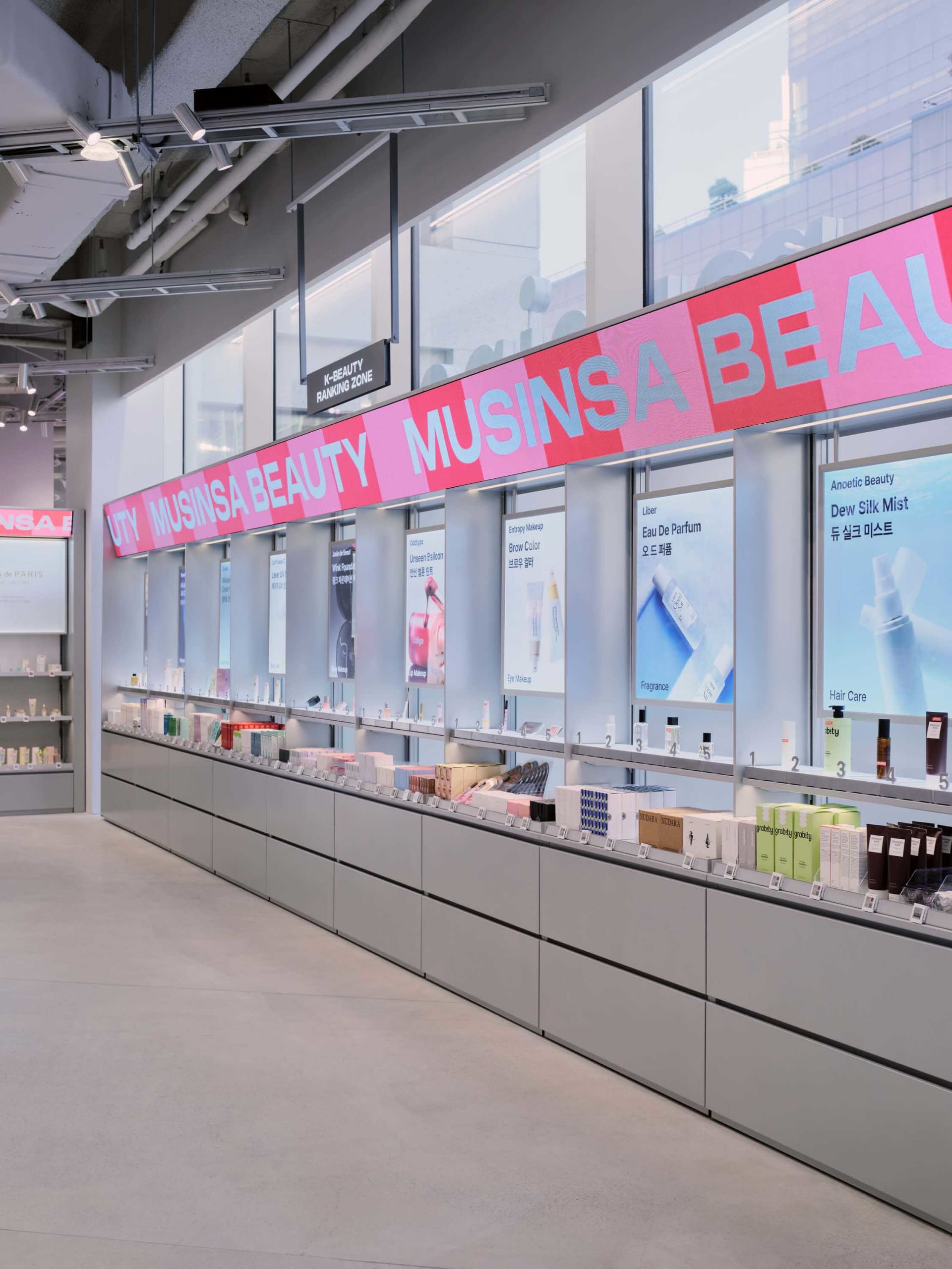 Long minimalist retail display wall in a modern beauty store with grey cabinetry, backlit product niches, and large pink signage reading MUSINSA BEAUTY.