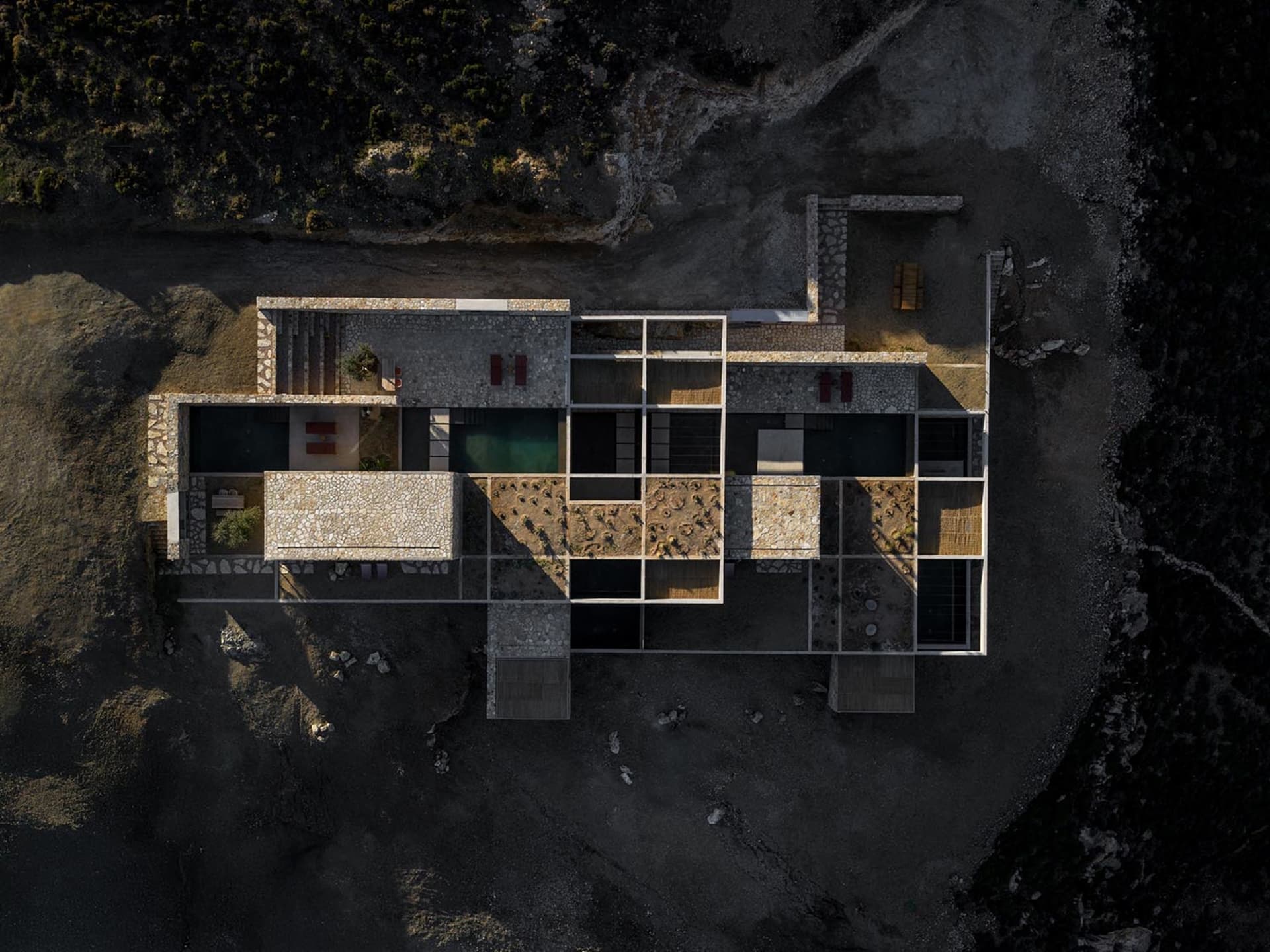 Aerial photograph of a contemporary stone courtyard villa featuring multiple infinity pools, open-air living spaces, and interconnected stone pavilions set in a rocky, arid terrain.