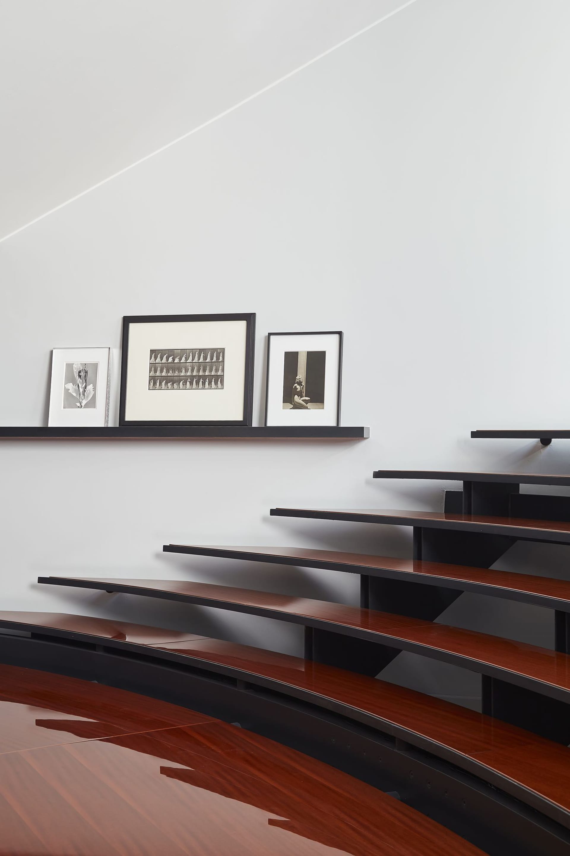 Modern curved walnut wooden staircase with glossy finish, black floating shelves displaying framed black-and-white photographs against a white wall.