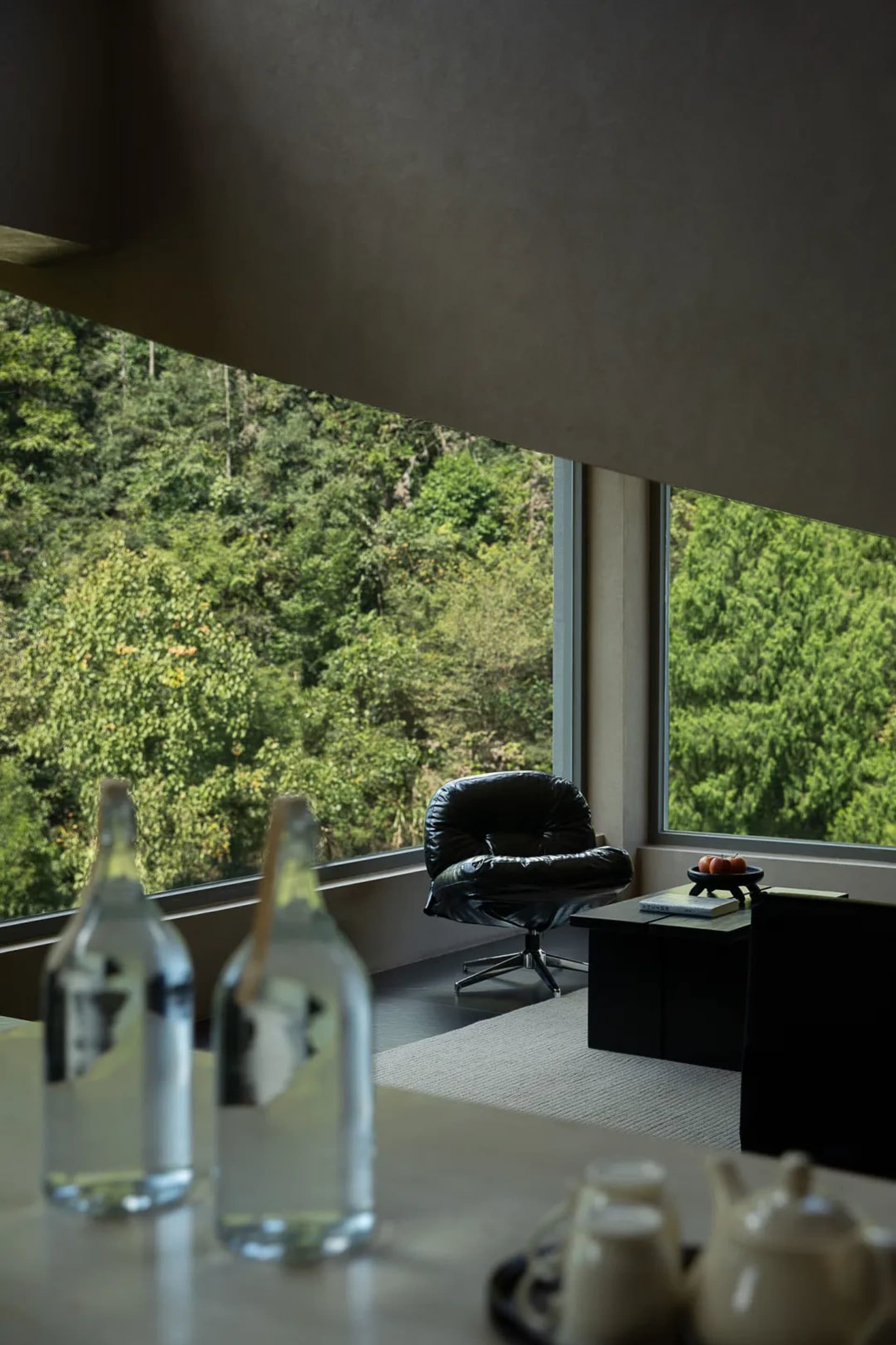 Modern living room interior with a black leather lounge chair placed beside floor-to-ceiling windows overlooking a dense green forest, with a low black side table and foreground table setting.