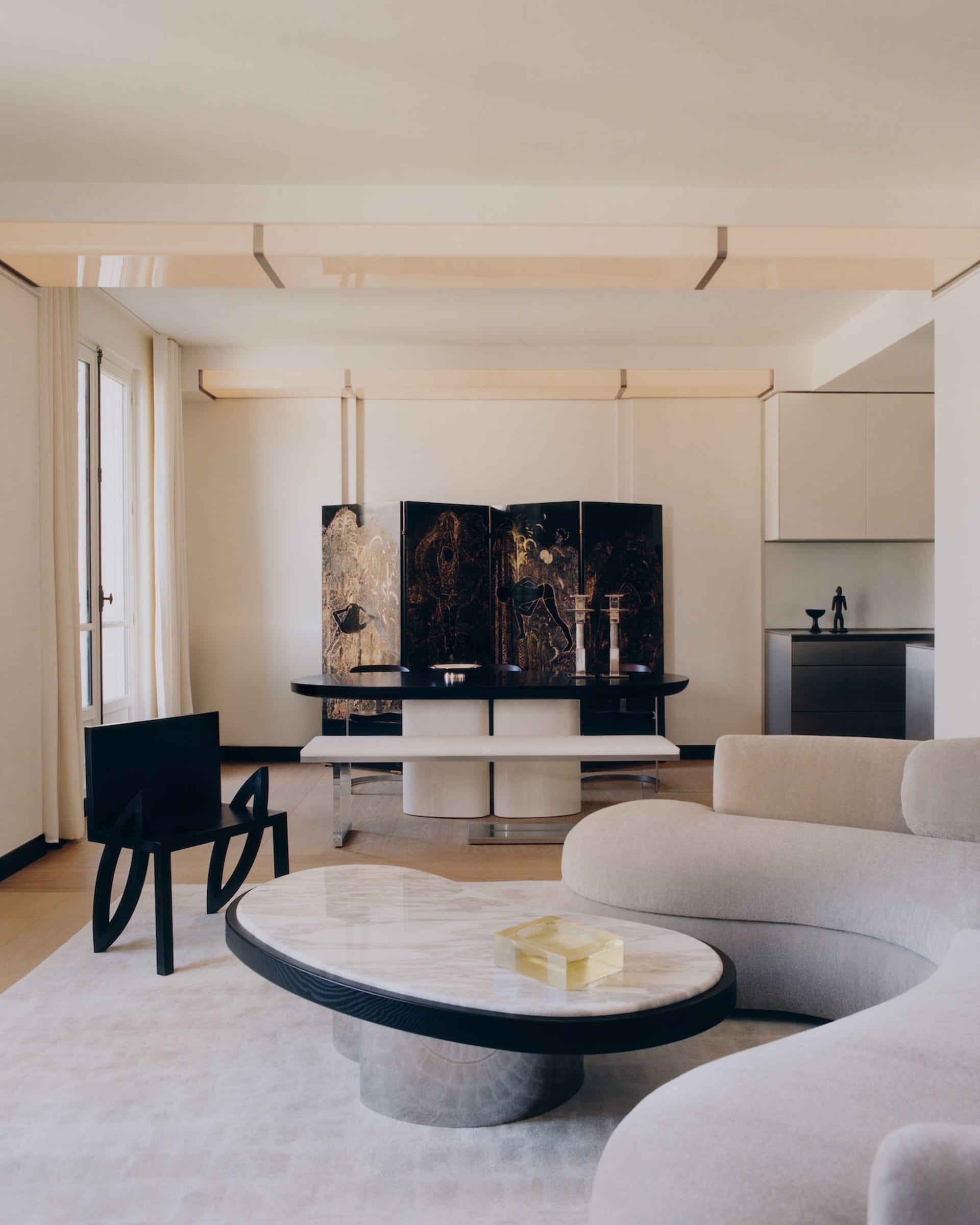 Modern minimalist living and dining area featuring a curved white marble coffee table, cream bouclé sectional sofa, black sculptural chair, and a large four-panel black lacquered folding screen with gold artwork.