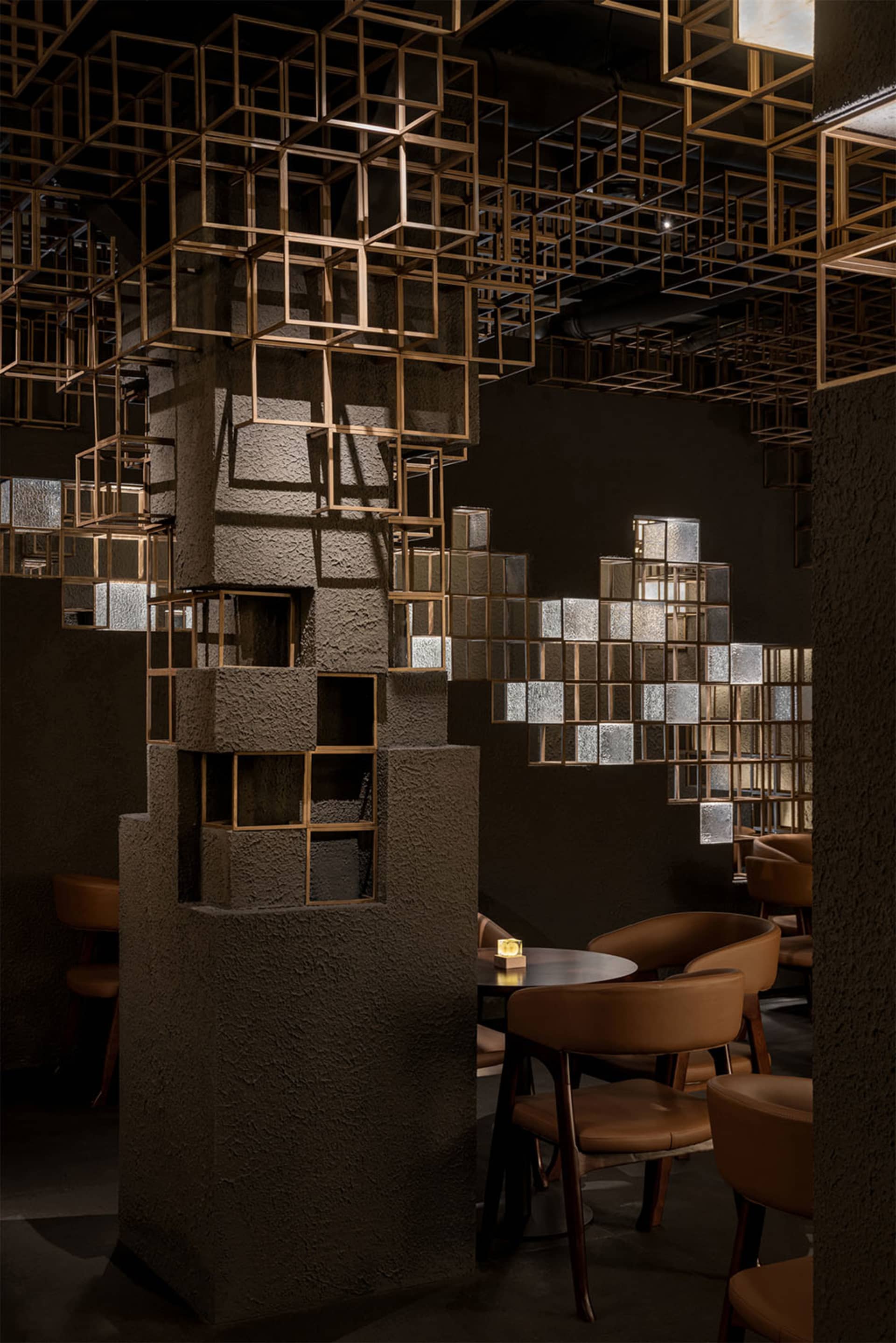 Interior of a stylish restaurant featuring textured concrete columns, brown leather dining chairs, round tables, and an elaborate ceiling installation of illuminated golden cubic metal frames.