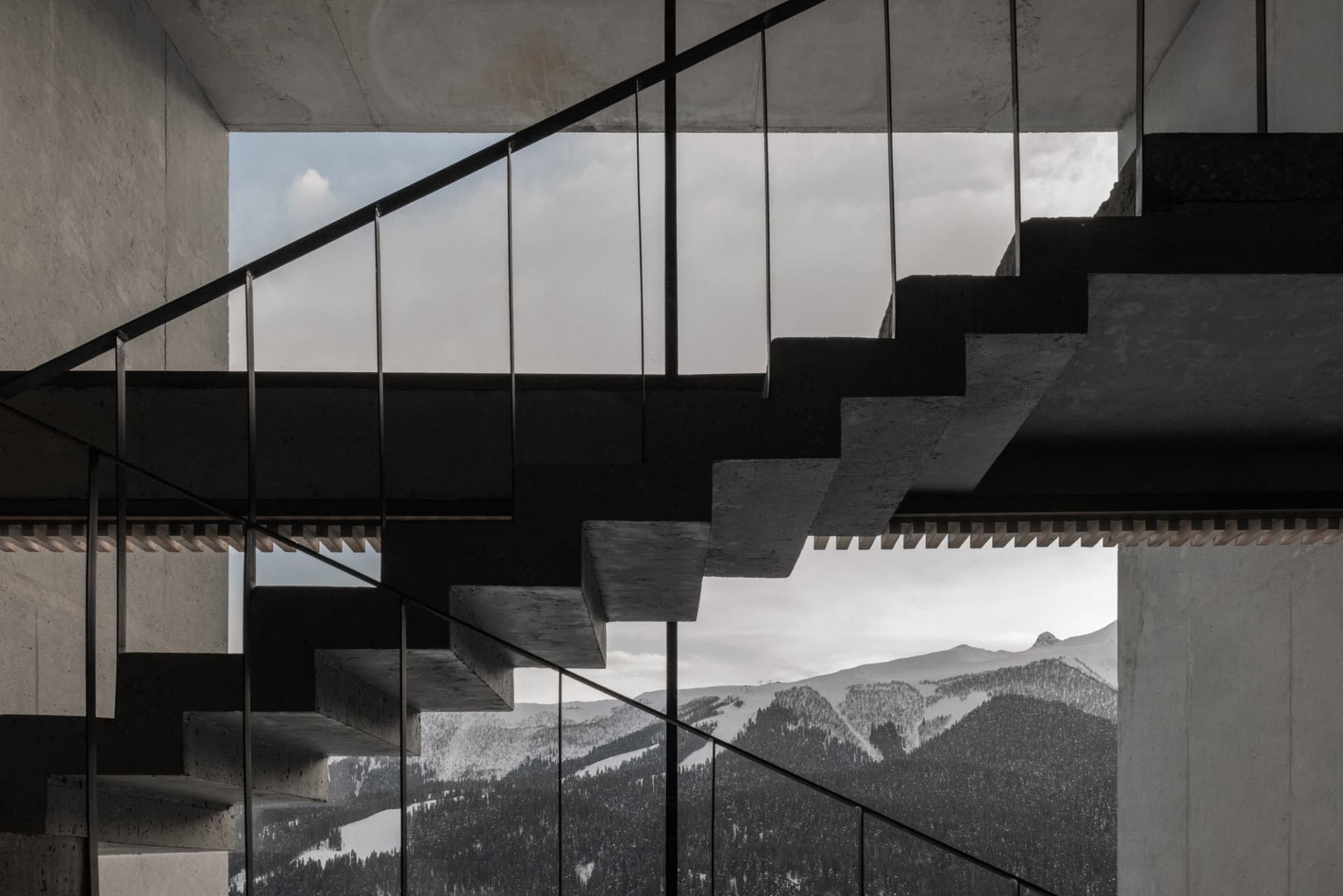 Modern concrete staircase with black metal railings overlooking snow-capped mountains and forested valleys.