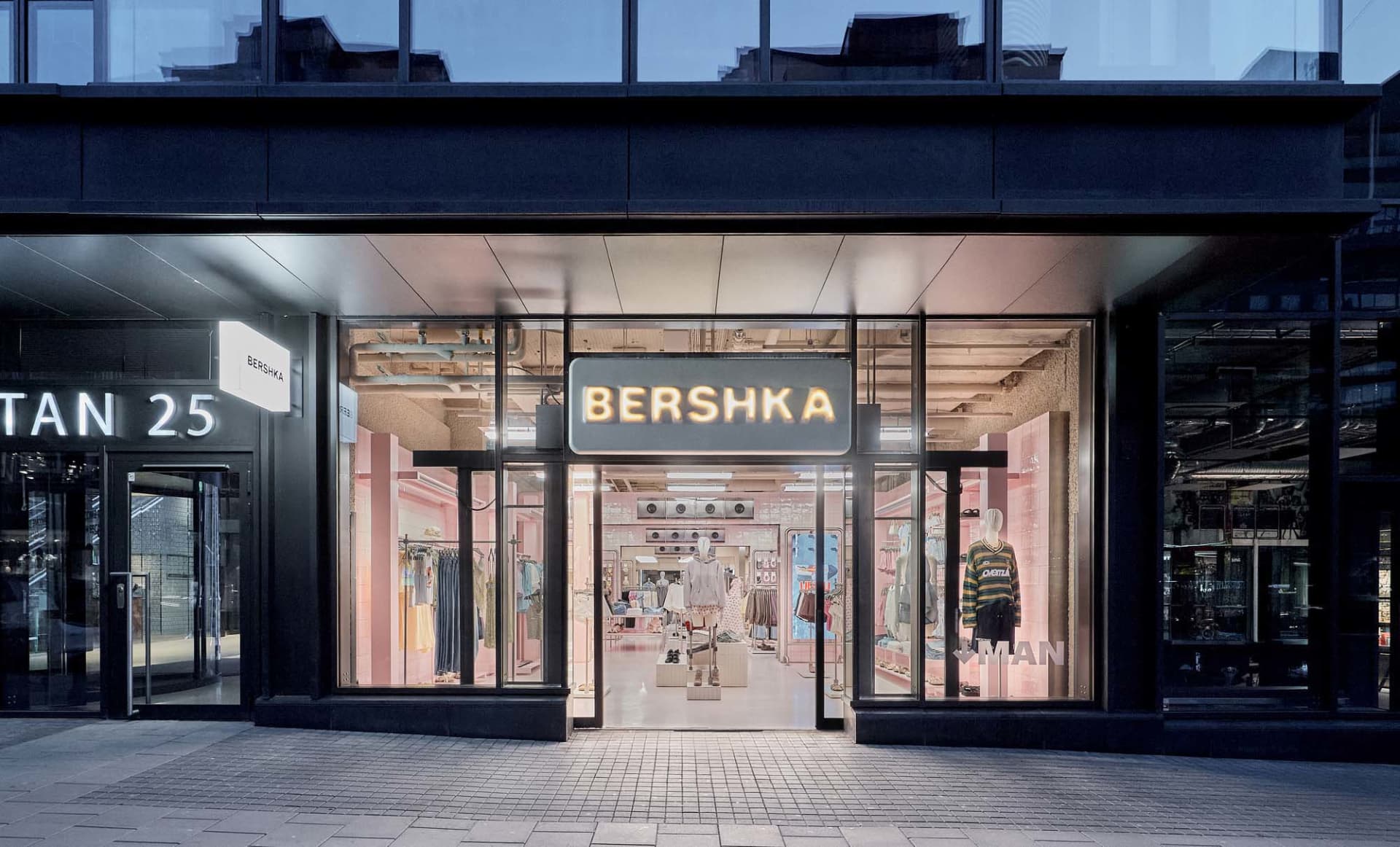 Night view of a modern Bershka retail store with large glass windows, pink interior lighting, visible clothing displays and mannequins, and illuminated signage.