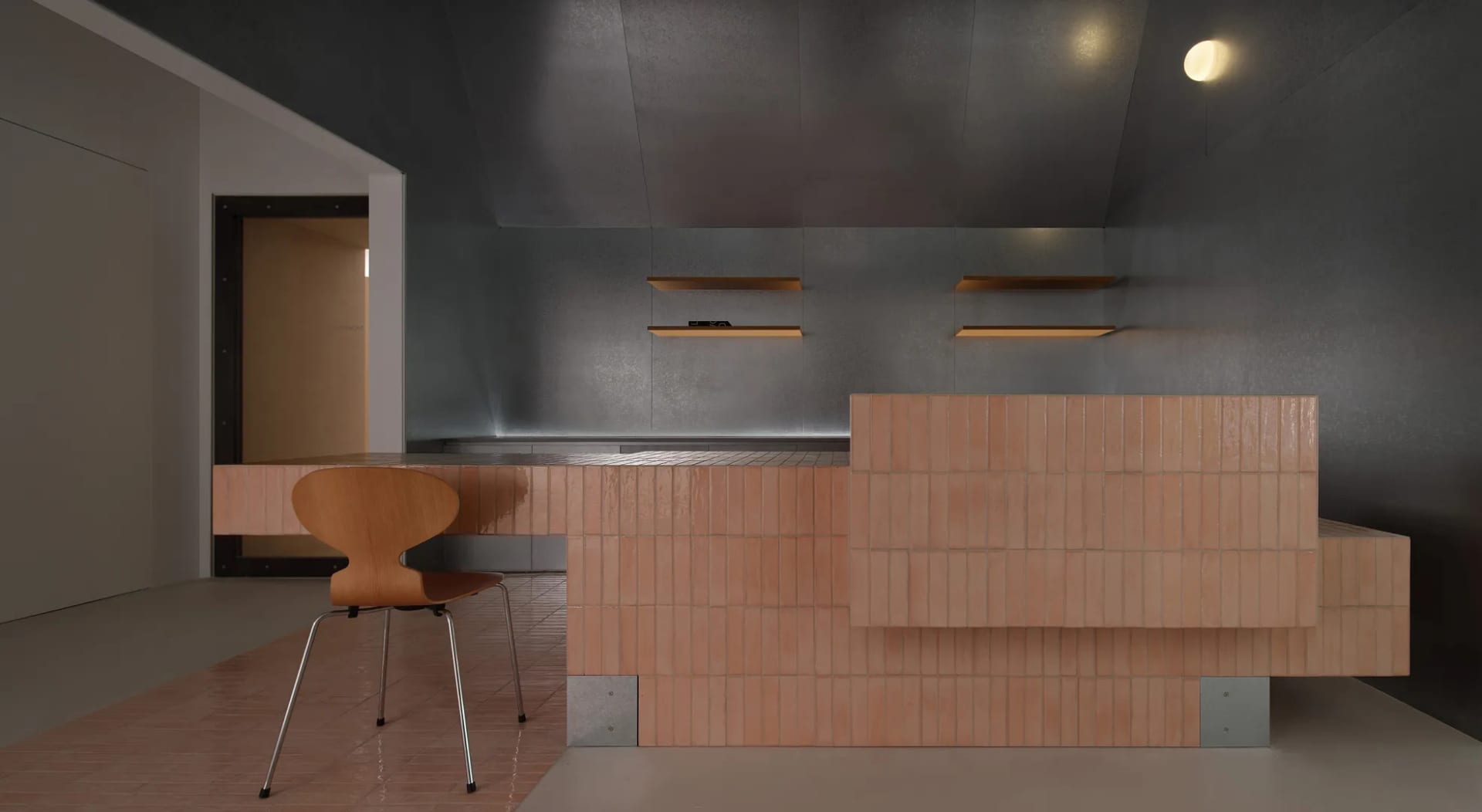 Modern minimalist kitchen featuring a terracotta tiled island counter, raw concrete walls and ceiling, wooden chair, and integrated warm lighting.