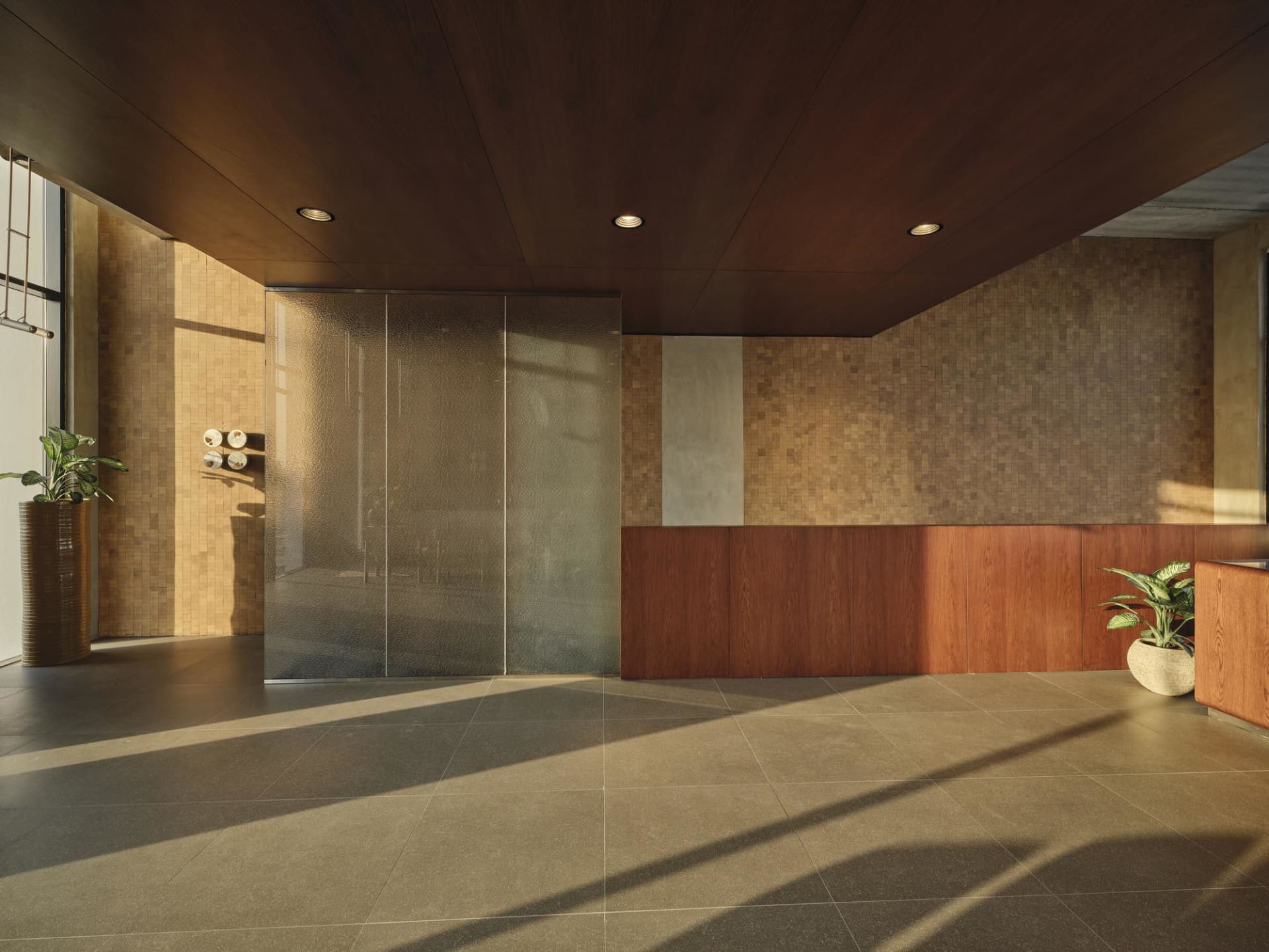 Modern commercial reception area with dark wood ceiling, warm wood paneling, beige mosaic tile walls, polished concrete floor, and potted plants.