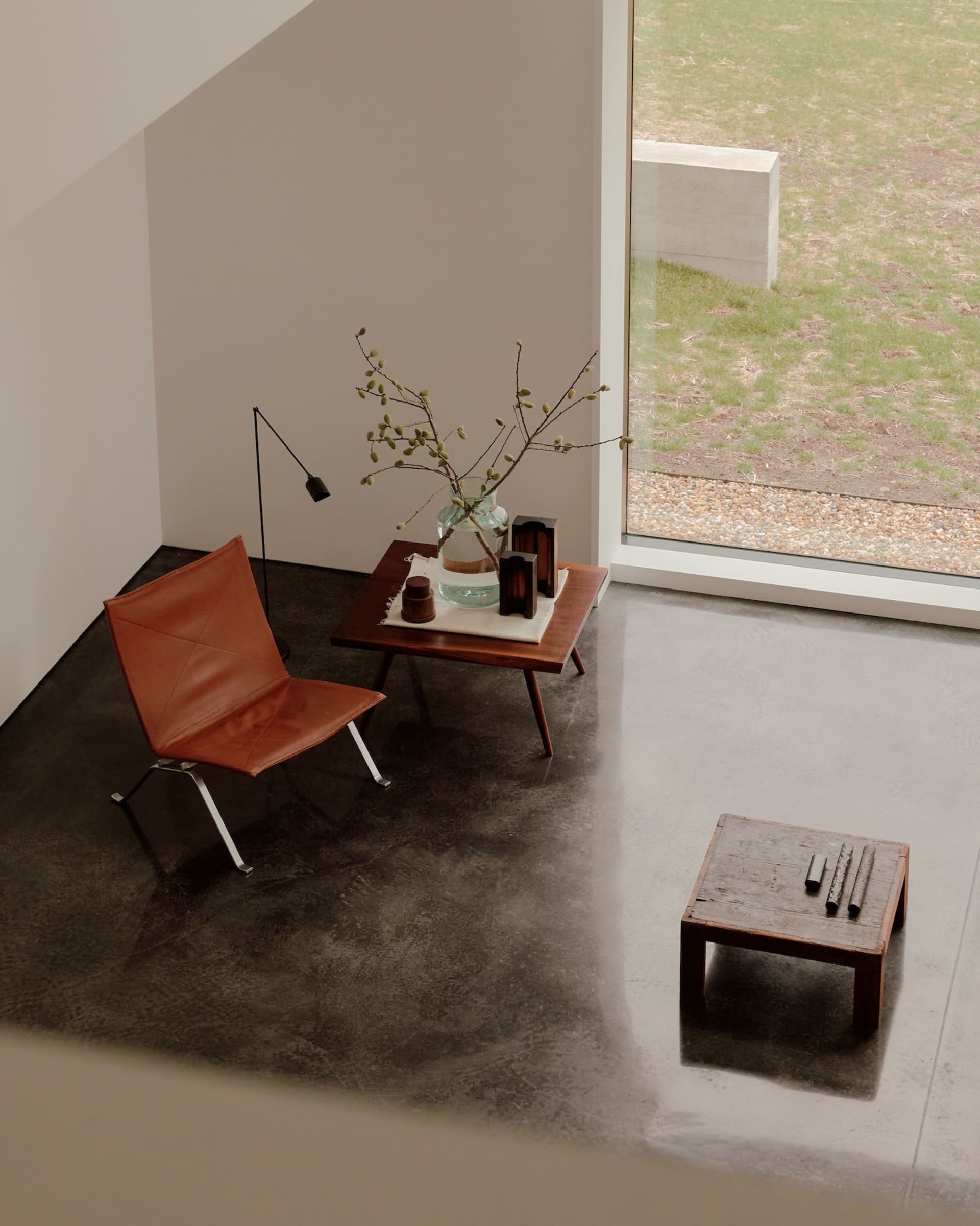 Modern minimalist interior featuring a brown leather sling chair, dark walnut side table with glass vase and branches, black floor lamp, and polished concrete floor next to a large window overlooking a green garden.