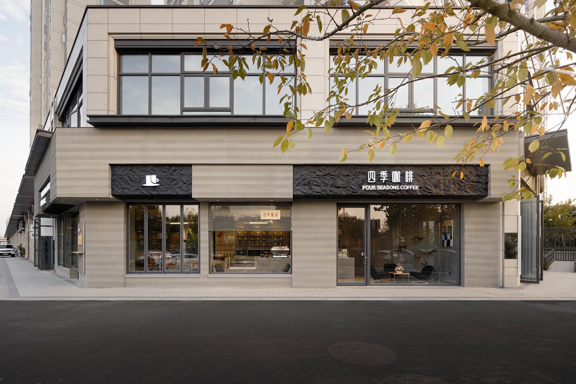 Modern coffee shop exterior with light travertine stone facade, large glass windows, and black textured stone signage reading Four Seasons Coffee.