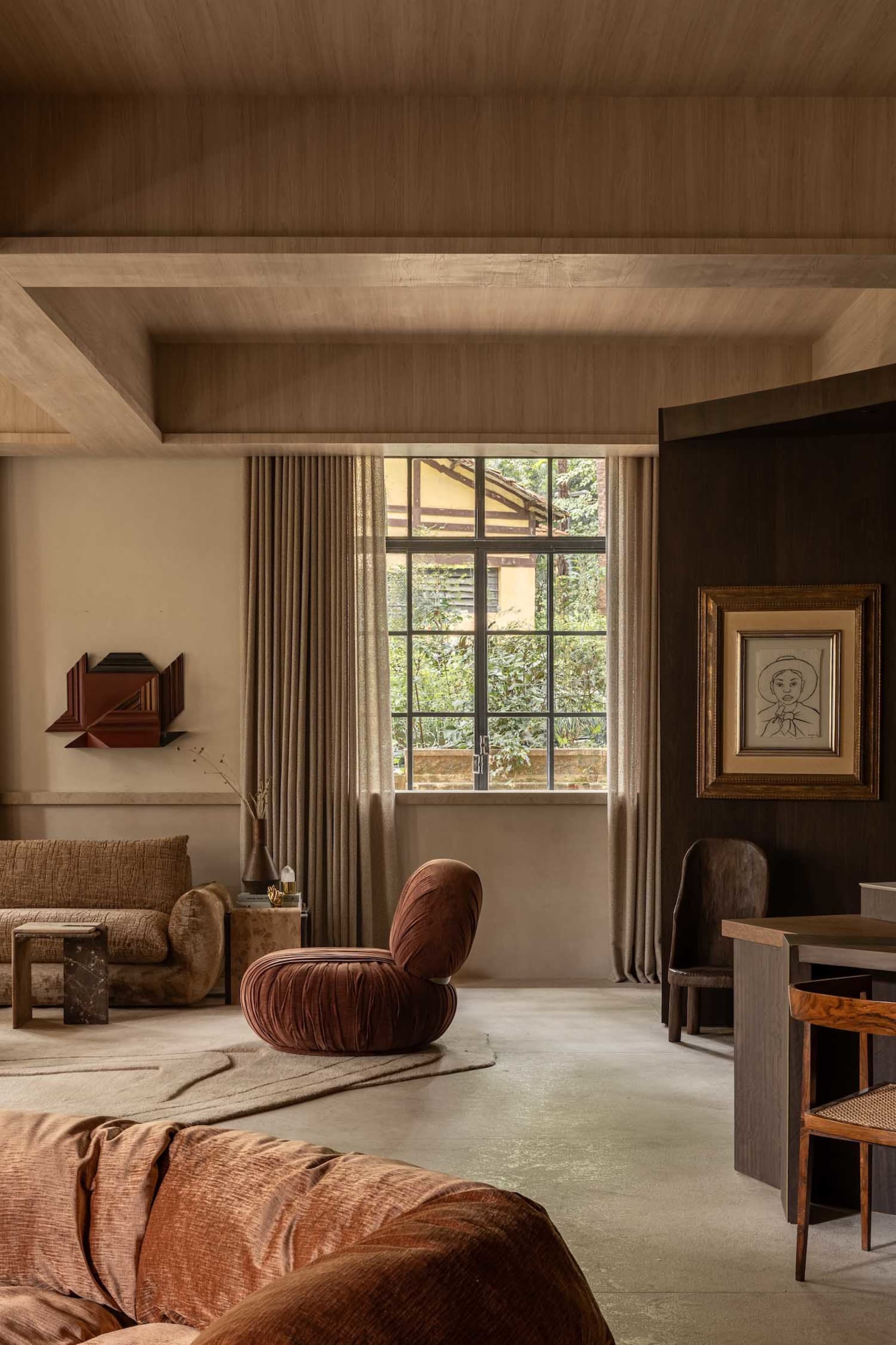 Modern living room with terracotta velvet lounge chair, warm wood ceiling, neutral sofa, large window with greenery view, and framed line drawing art.
