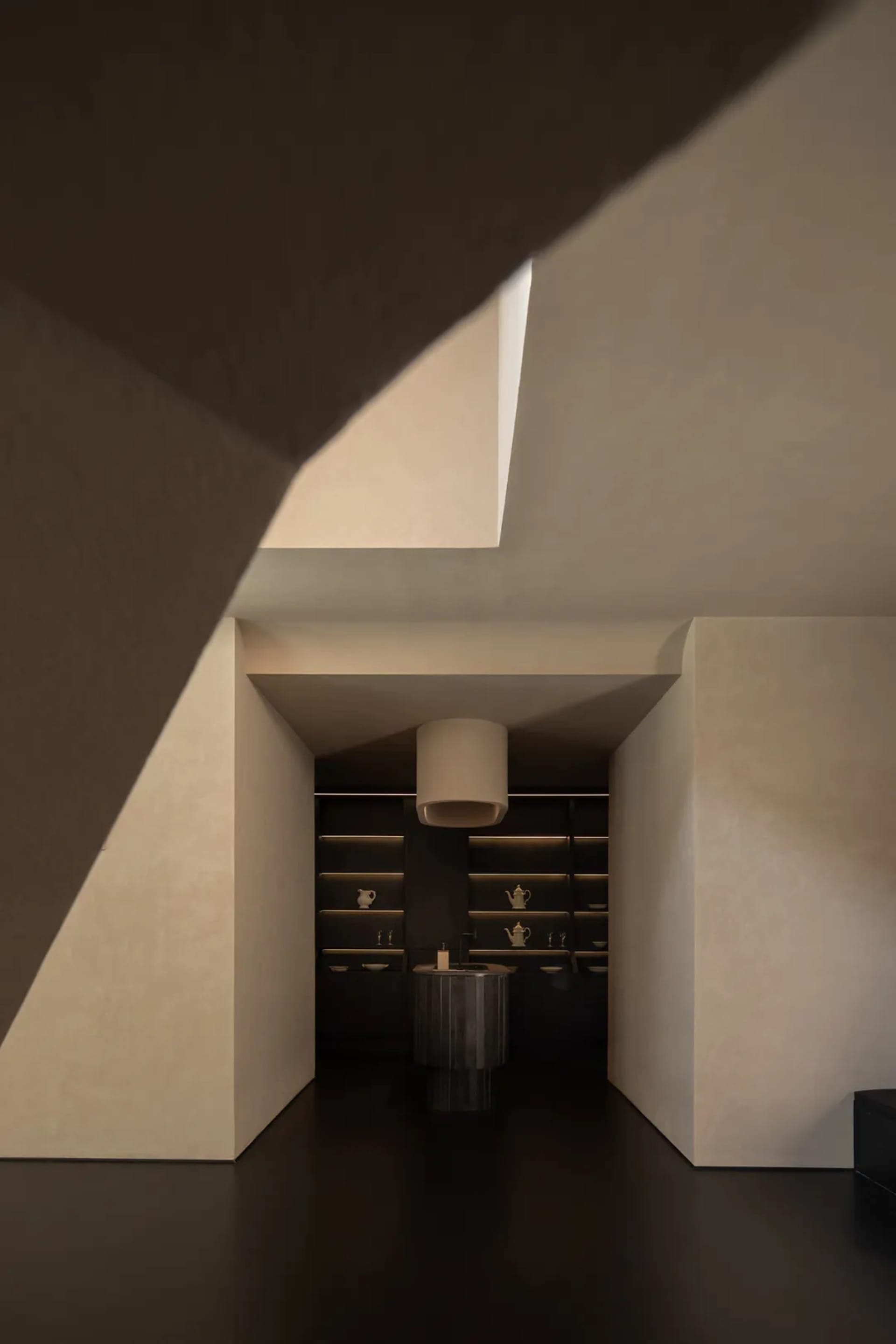 Modern minimalist butler pantry viewed through a dramatic angular concrete opening, featuring dark micro-cement walls, black fluted island, and illuminated open shelving.