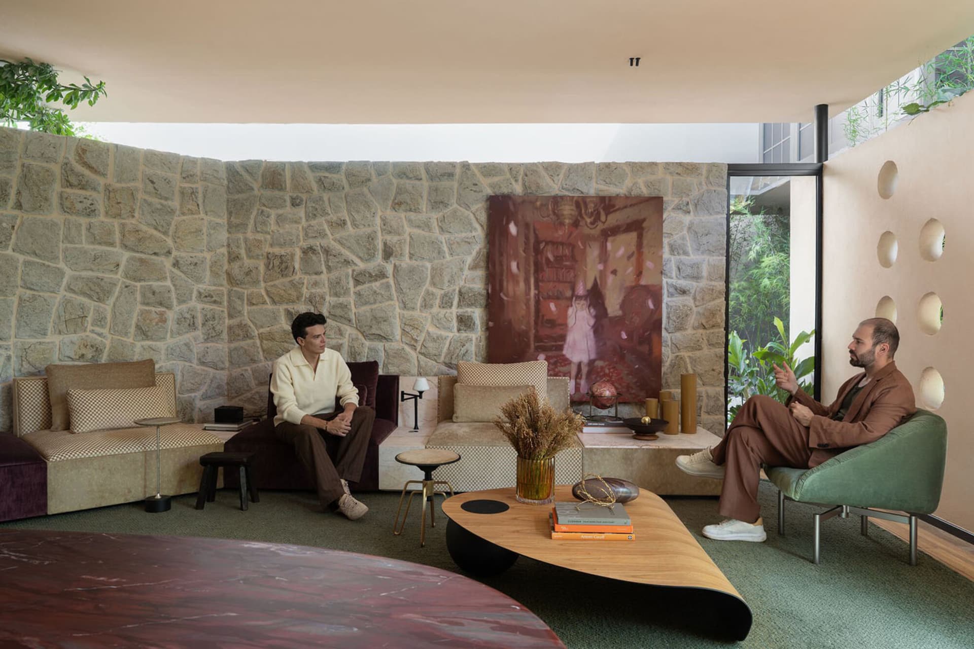 Modern living room featuring a dramatic curved natural stone wall, mixed burgundy and beige velvet sofas, a green swivel armchair, and an organic-shaped wooden coffee table with a large abstract painting in the background.