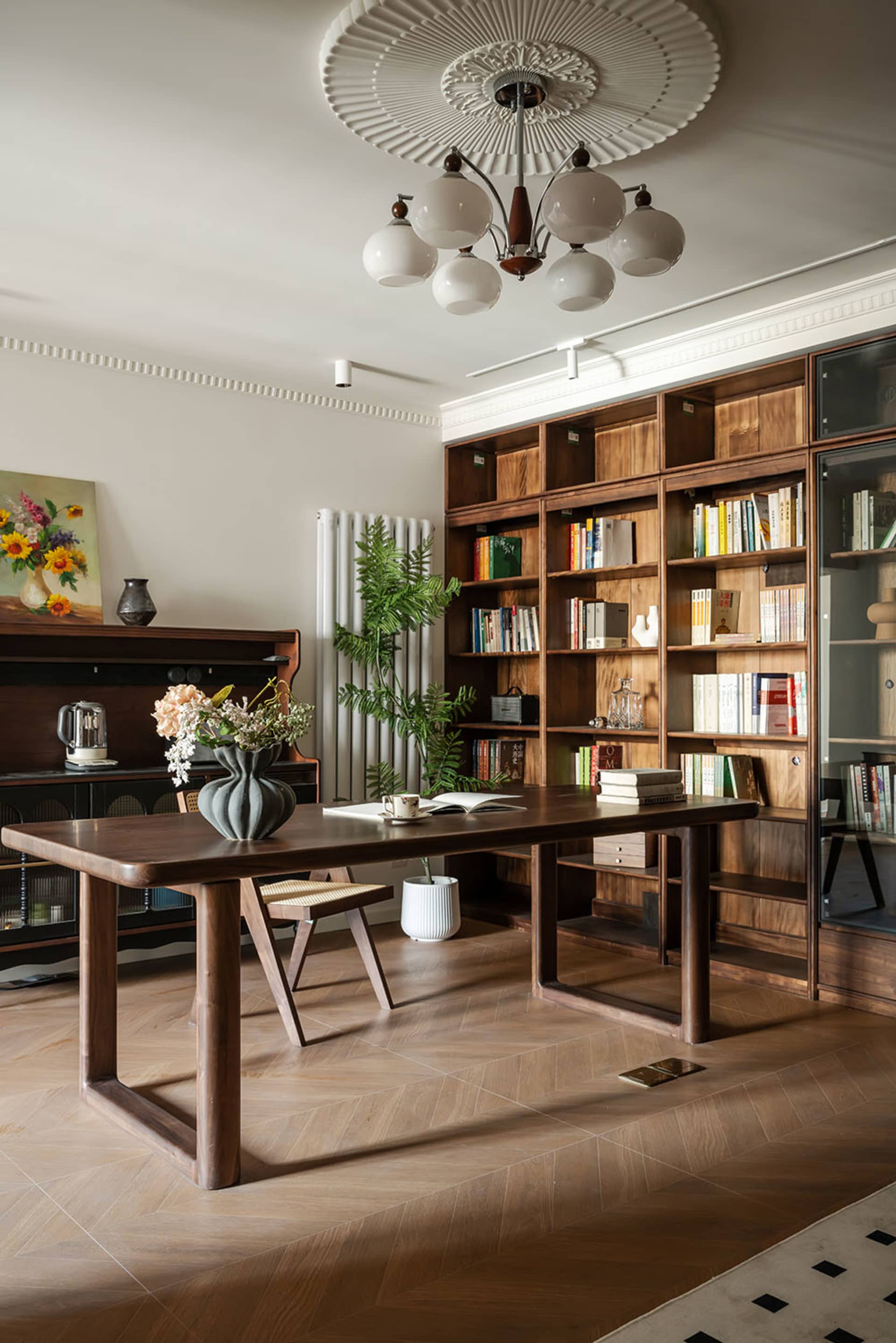 Spacious home library with floor-to-ceiling walnut built-in bookshelves, a large walnut writing desk, woven stool, potted plants, and an ornate white ceiling medallion with a modern chandelier.