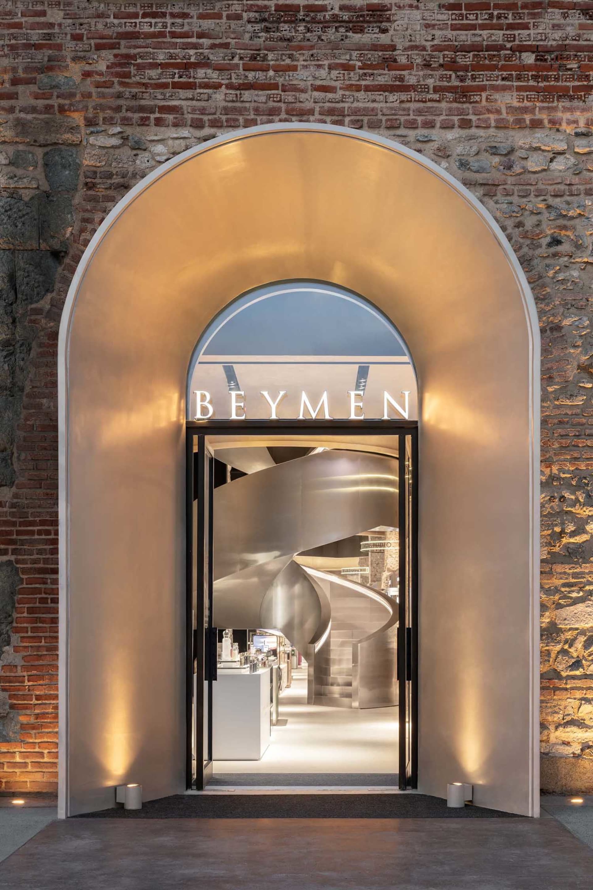 View of a modern luxury store entrance featuring a large illuminated stainless steel arch set within an old brick wall, revealing a dramatic spiral staircase inside.