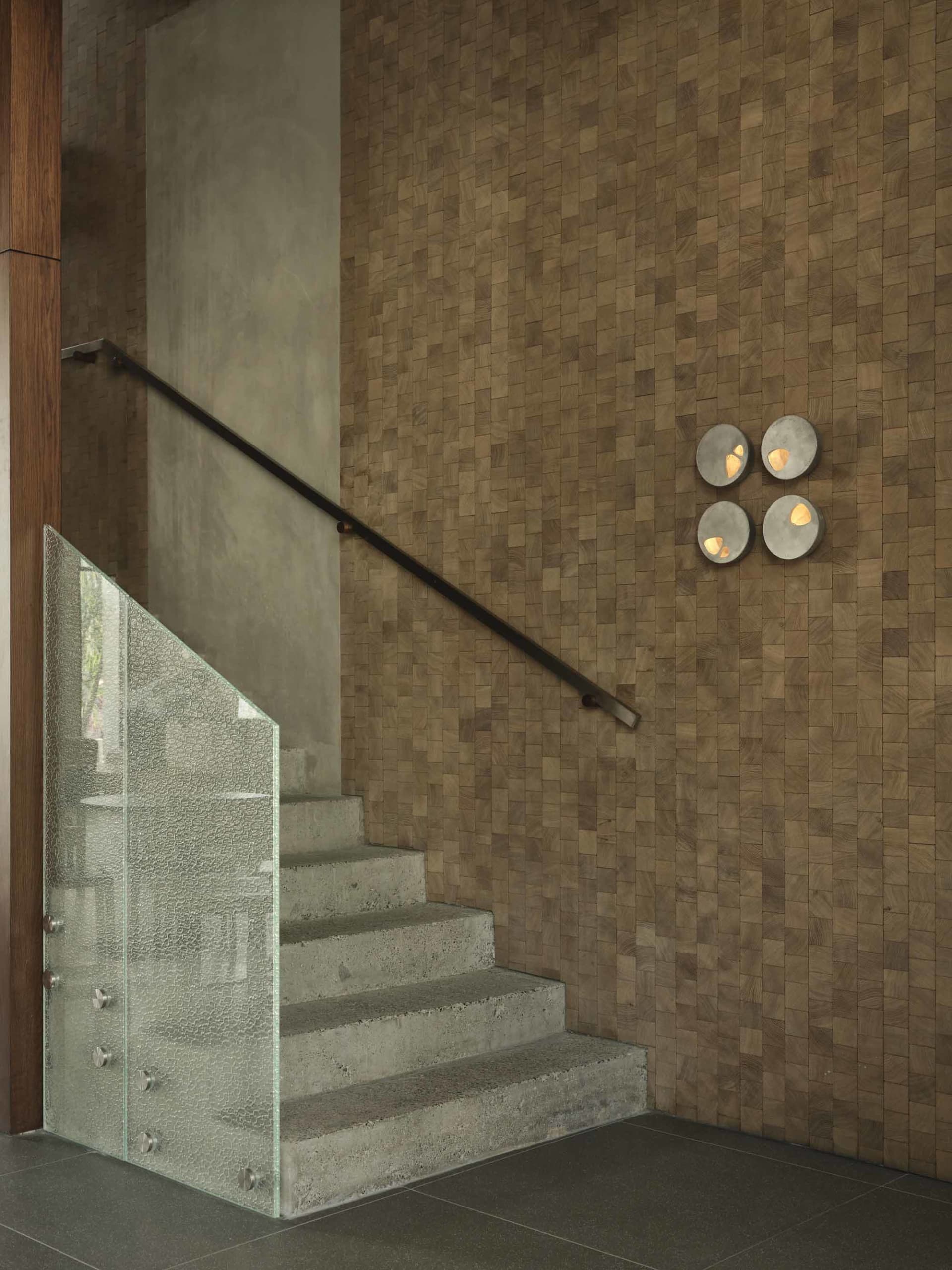 Modern concrete staircase with textured glass railing beside a wall covered in small wooden blocks and four circular wall sconces.