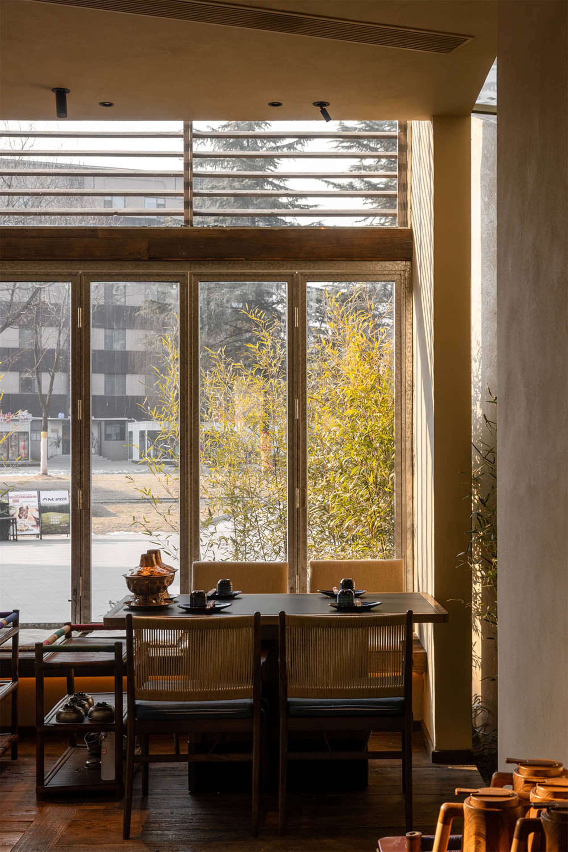 Modern Chinese-style dining room featuring dark wood table, woven rattan dining chairs, warm wooden flooring, and large glass doors opening to a lush bamboo garden with natural daylight.