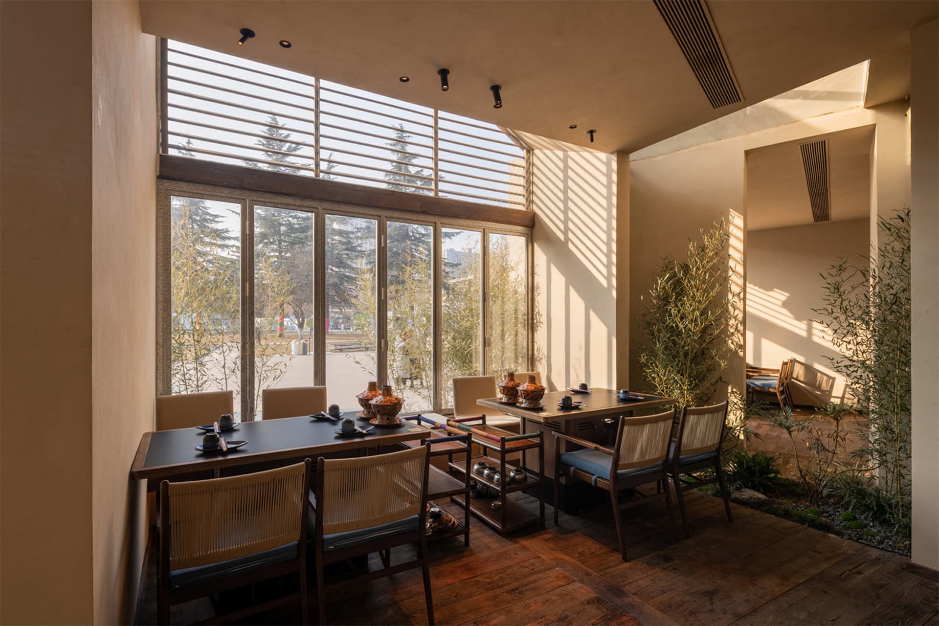 A modern Japanese-style dining room featuring dark wood tables, woven rattan chairs with blue cushions, large glass windows with wooden shutters overlooking bamboo and trees, and potted bamboo plants inside.