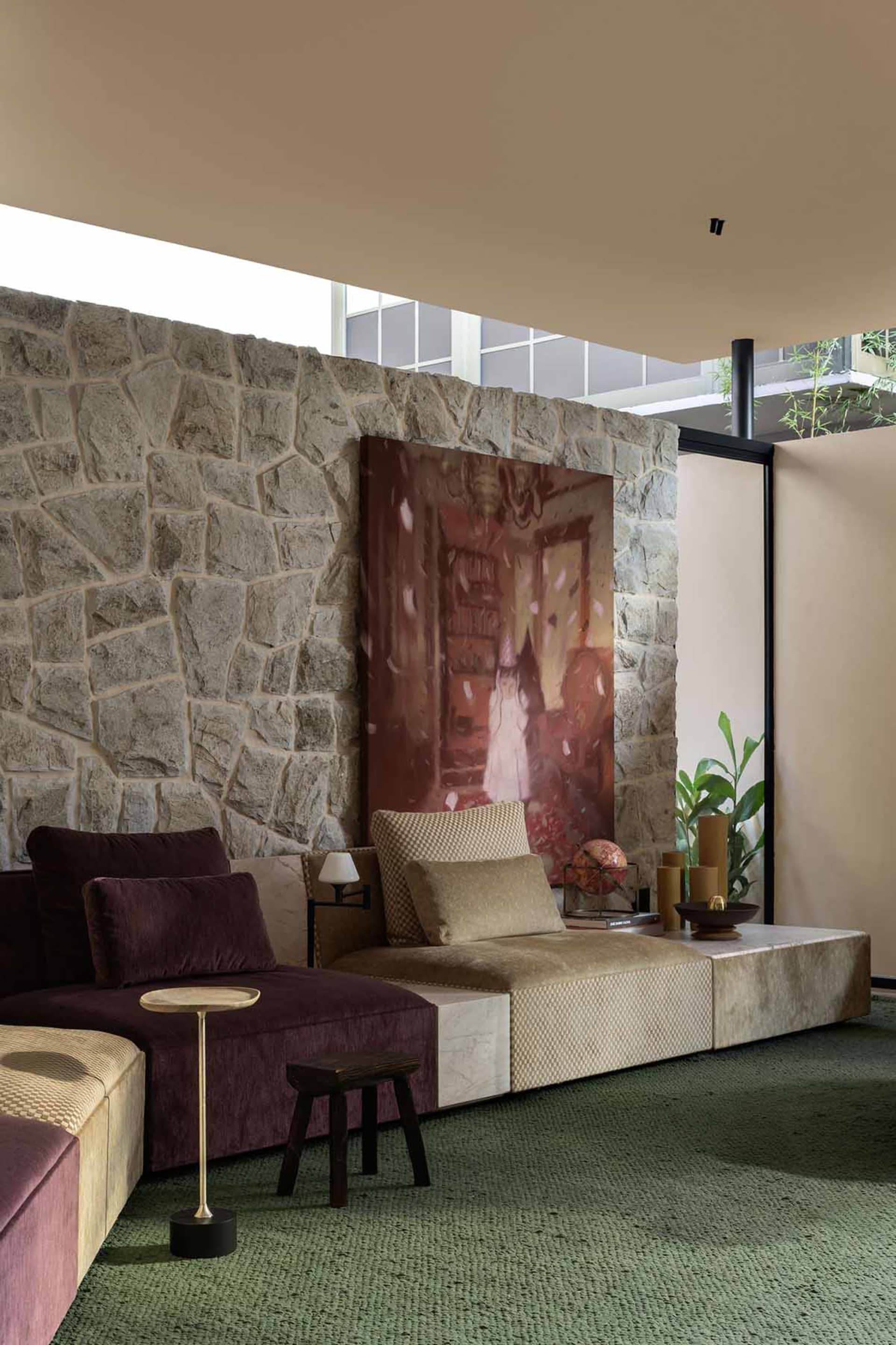 Modern lounge area featuring a large textured stone wall, a modular sofa with deep maroon velvet and beige bouclé sections, a substantial abstract painting, and green textured carpet.