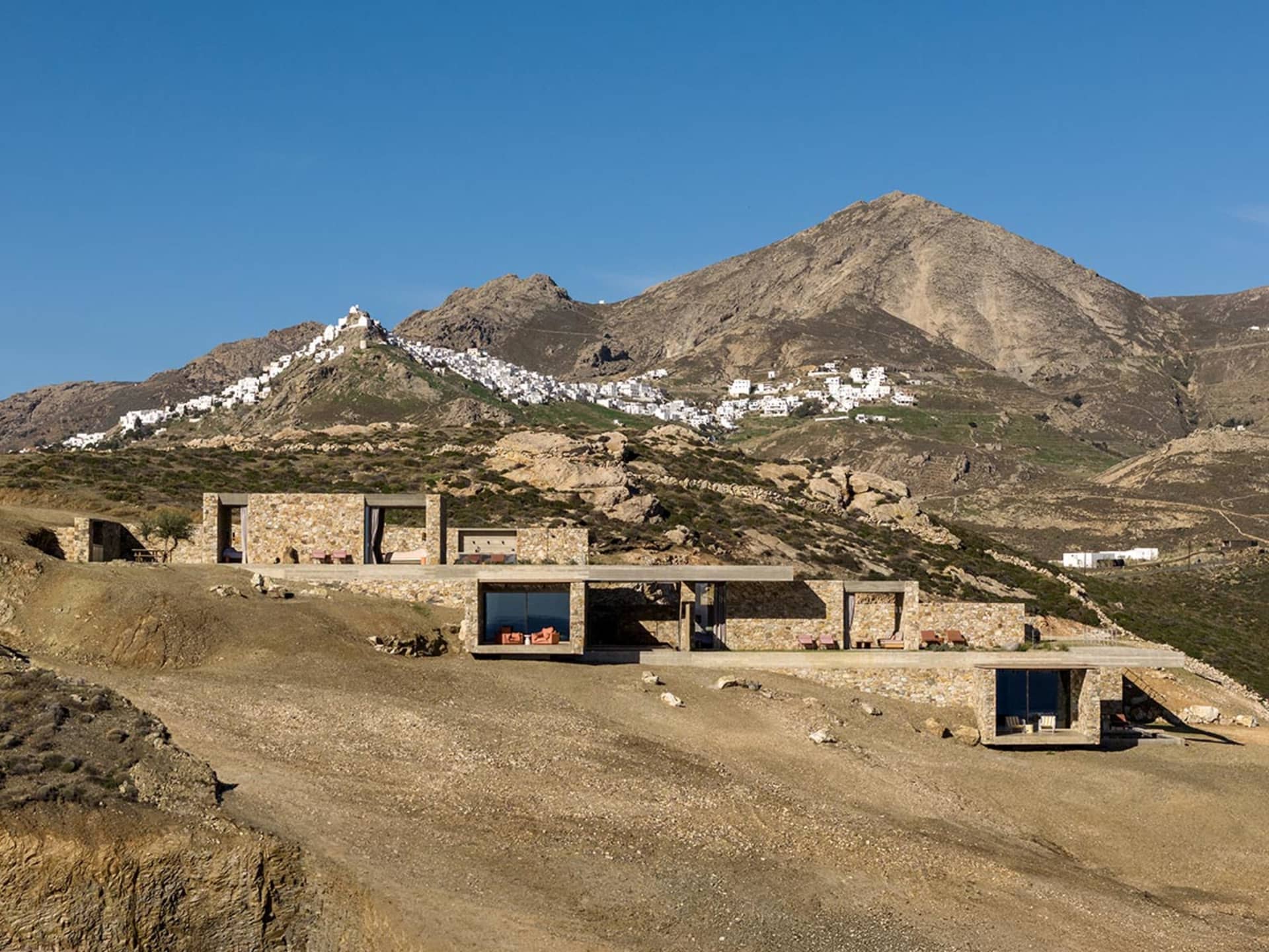 現代石材別墅擁有大型玻璃窗與戶外露台,建於乾燥山坡上,俯瞰希臘山上的白色希臘島村落