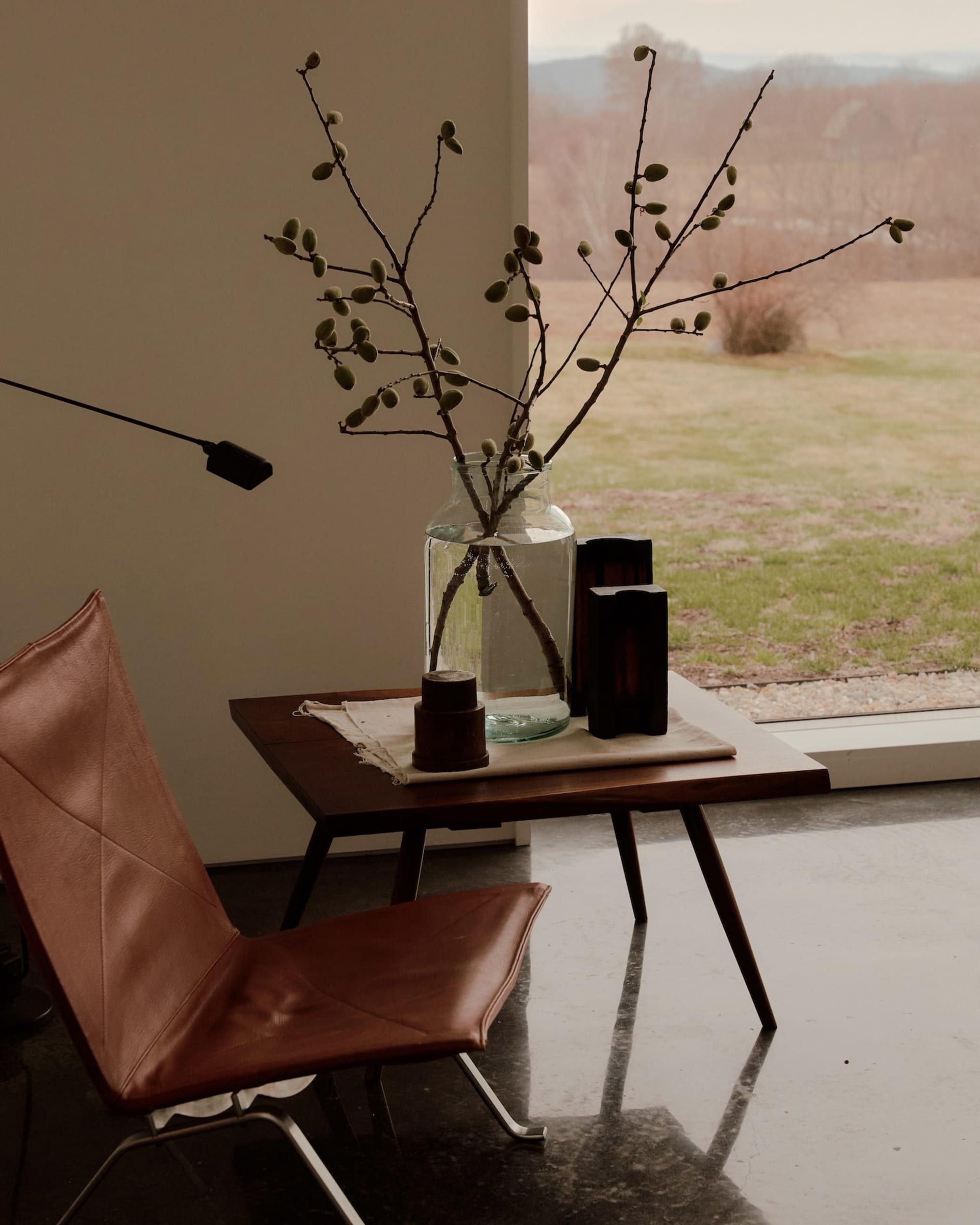 Cognac leather butterfly chair beside a walnut side table holding a large glass vase with budding branches, dark wooden objects, and a view of a winter landscape through a large window.