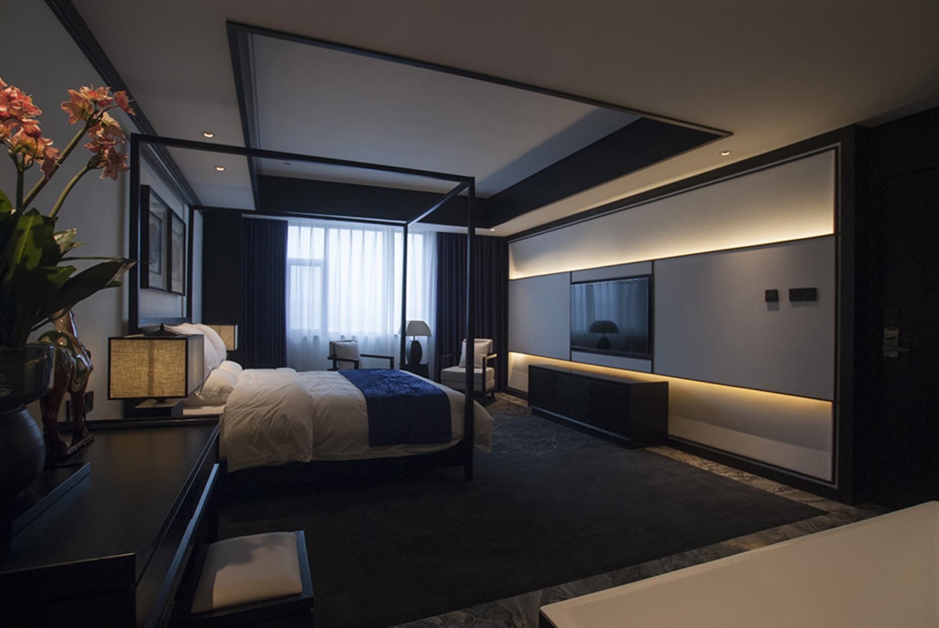 Modern luxury bedroom with black four-poster bed, dark wood furniture, blue throw, integrated wall-mounted TV, and layered ambient lighting.
