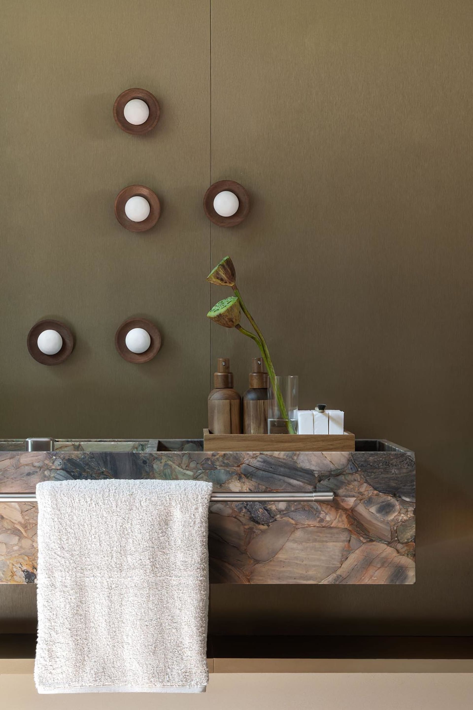 Modern bathroom featuring a floating vanity made of multicolored marble with an olive-green wall, wooden wall sconces, and a white towel hanging on a metal rod.