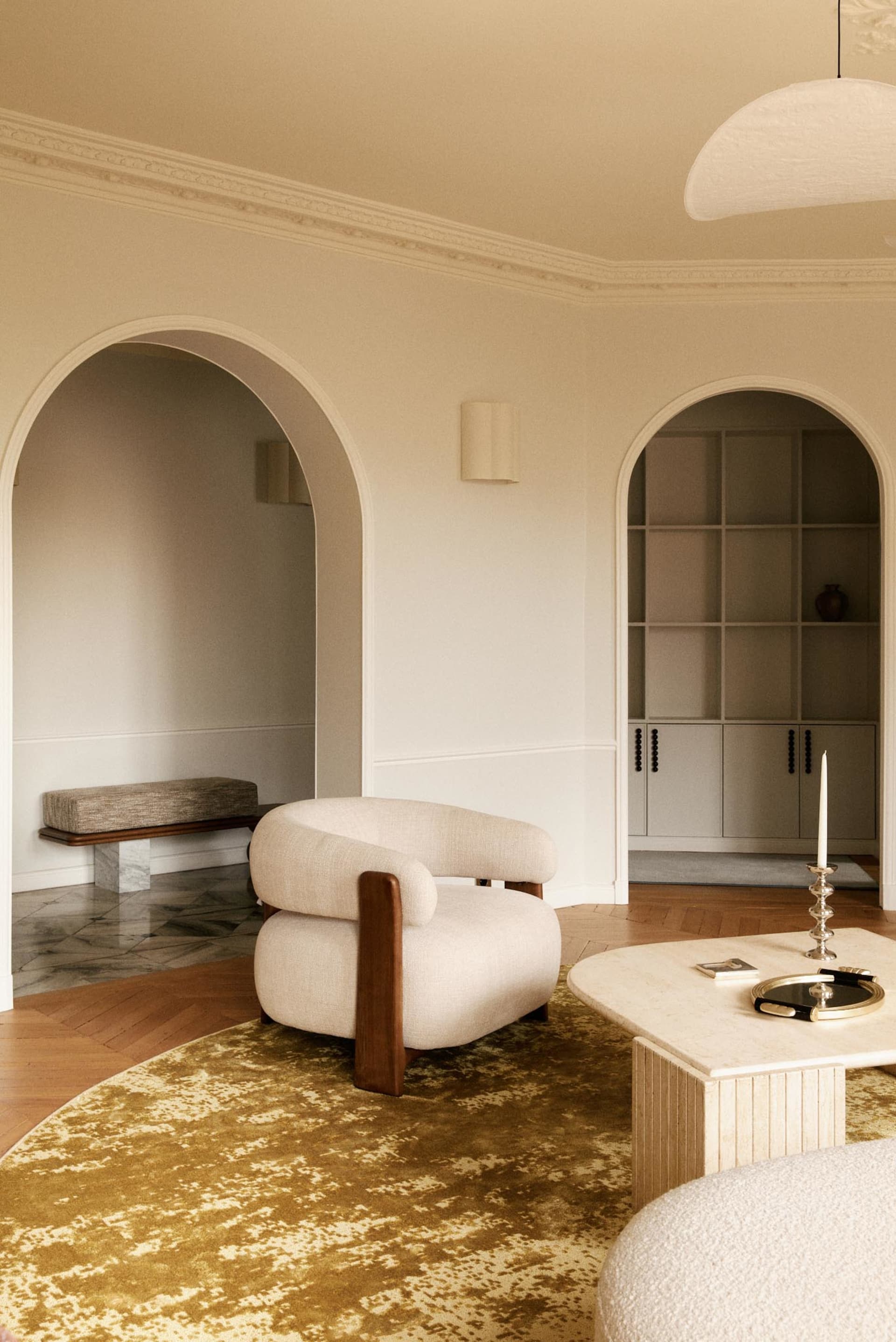 Modern living room featuring a curved cream linen armchair with walnut arms, a large travertine coffee table, mustard-yellow abstract rug, and multiple arched doorways leading to built-in shelving.
