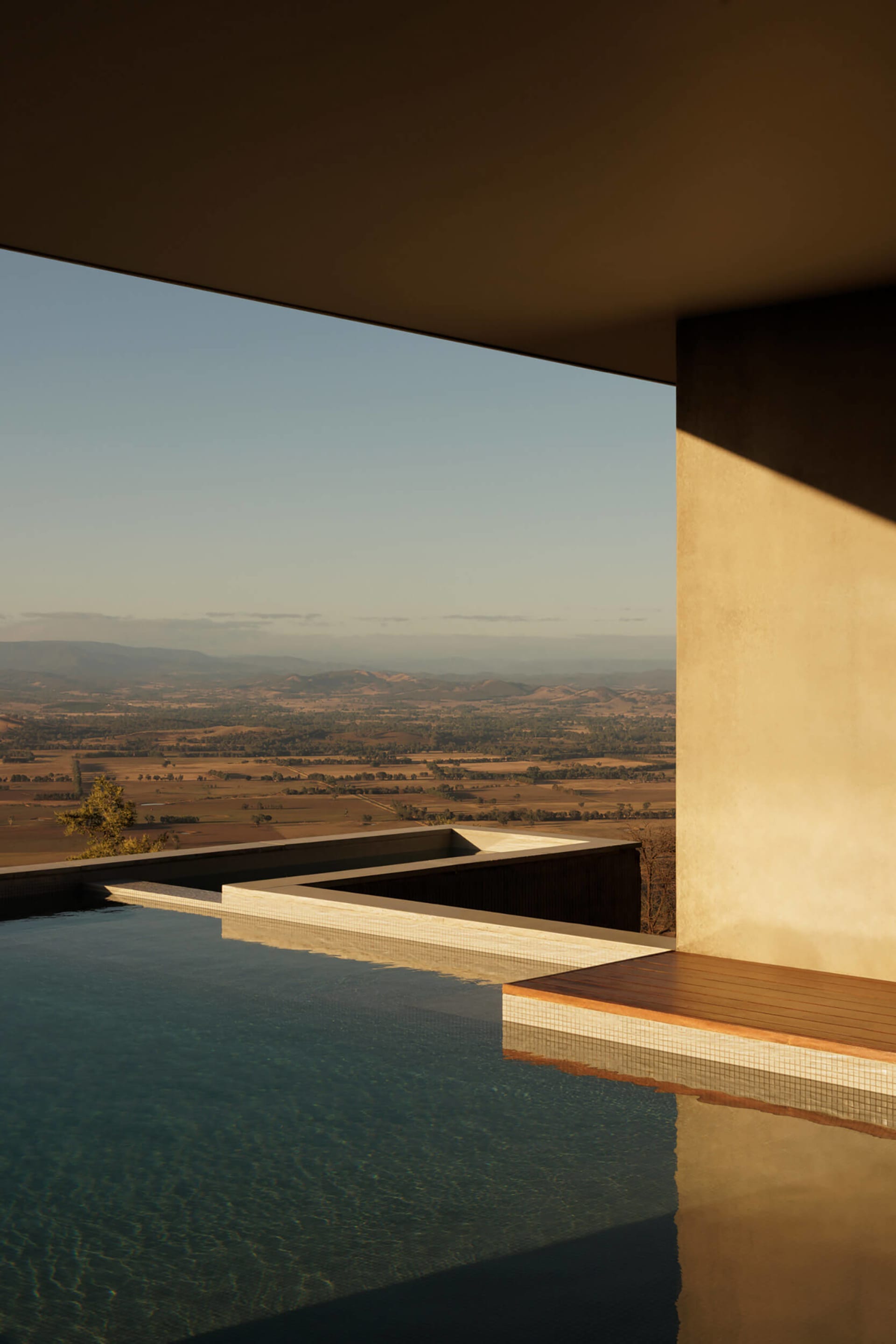 Modern infinity pool with dark water and concrete edge, set against a panoramic view of golden hills and farmland at sunset, framed by a minimalist concrete overhang and wooden deck.