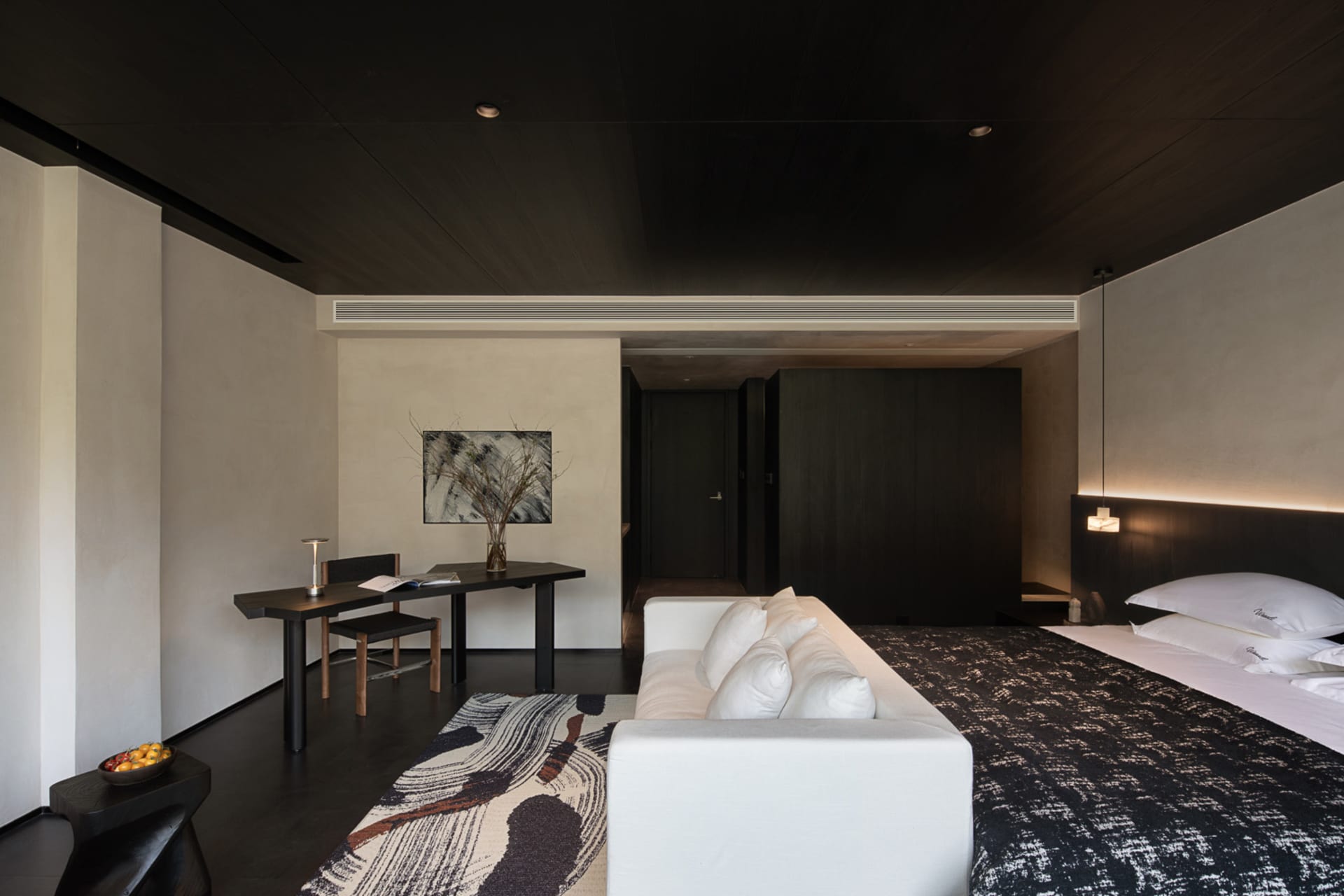 Modern minimalist bedroom featuring dark wood ceiling and walls, a large bed with white linens, a white sofa, an abstract black and white rug, and a built-in desk area with artwork.