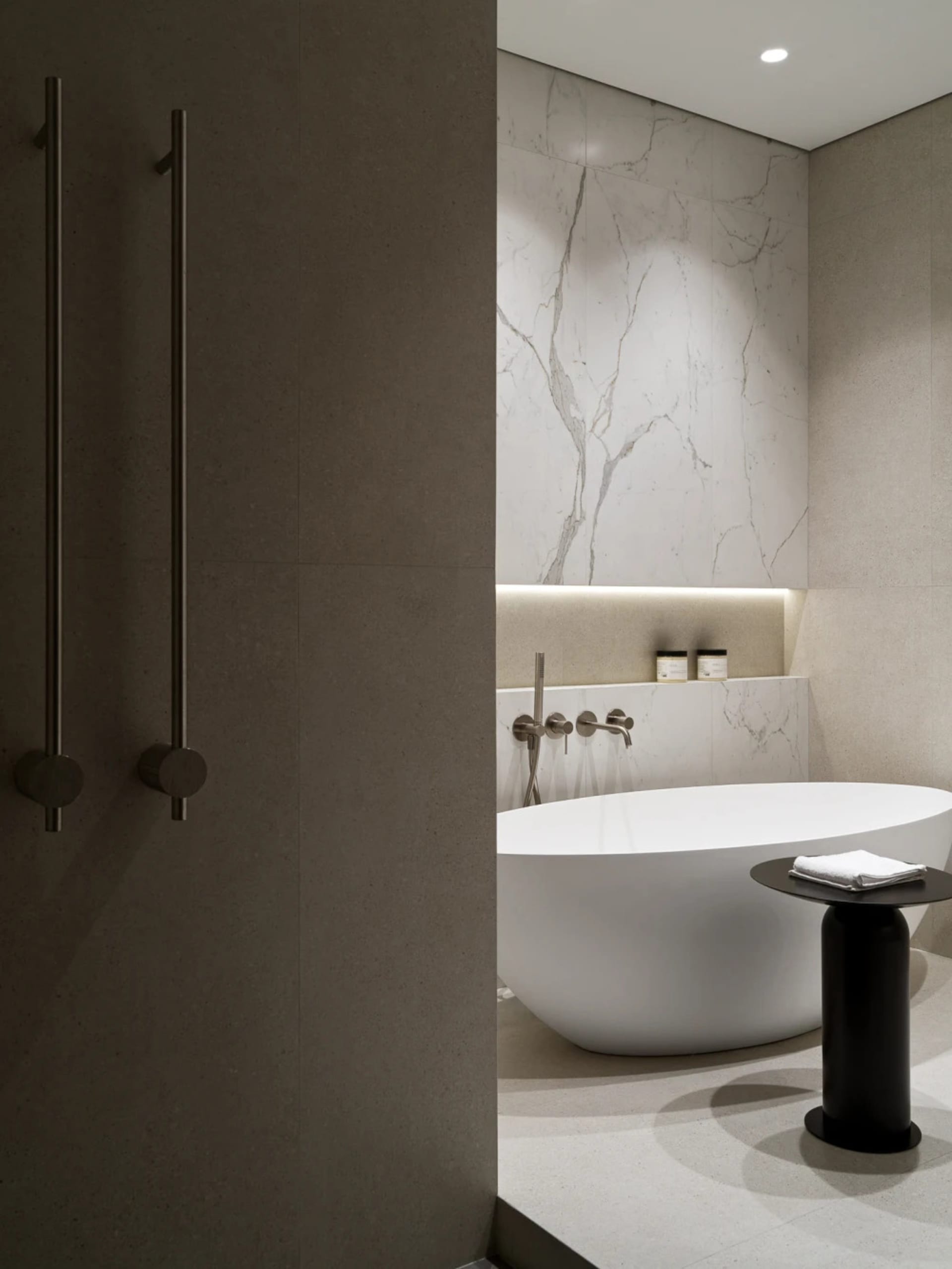 Modern minimalist bathroom featuring a white freestanding oval bathtub, large marble tile walls, recessed lighting, and a black cylindrical side table with folded white towels.