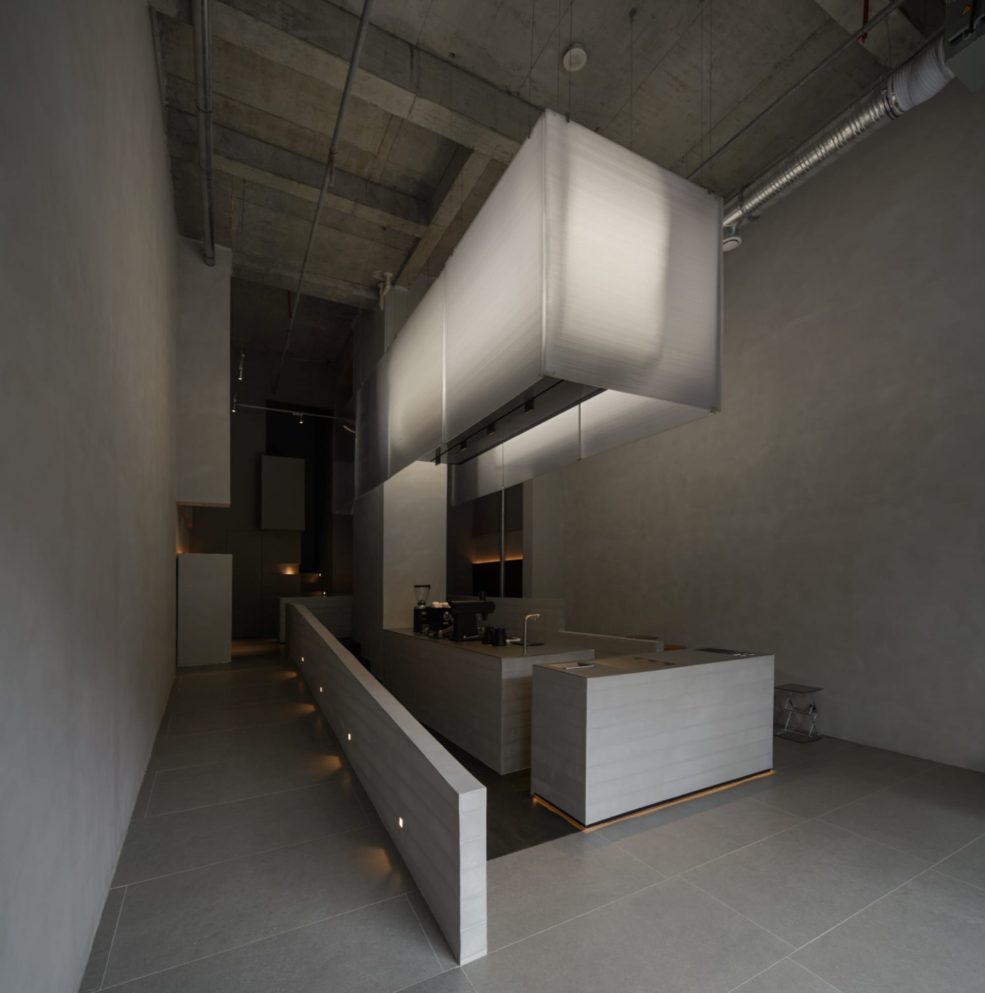 Modern brutalist commercial kitchen interior featuring a long concrete service counter, central kitchen island, and large rectangular fabric pendant lights suspended from an exposed concrete ceiling.