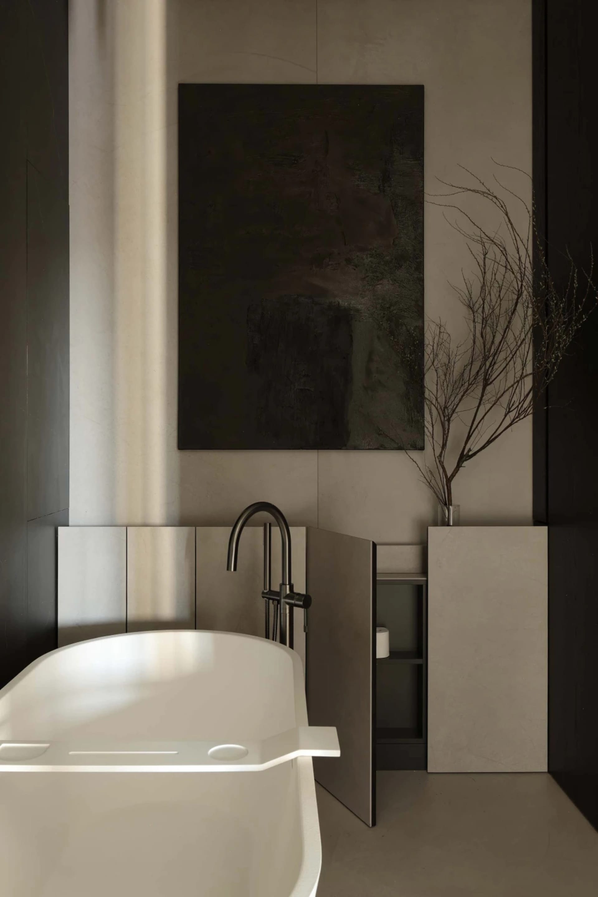 Modern minimalist bathroom featuring a white freestanding bathtub, matte concrete walls, black matte faucet, dark abstract painting, and dried branches in a glass vase.