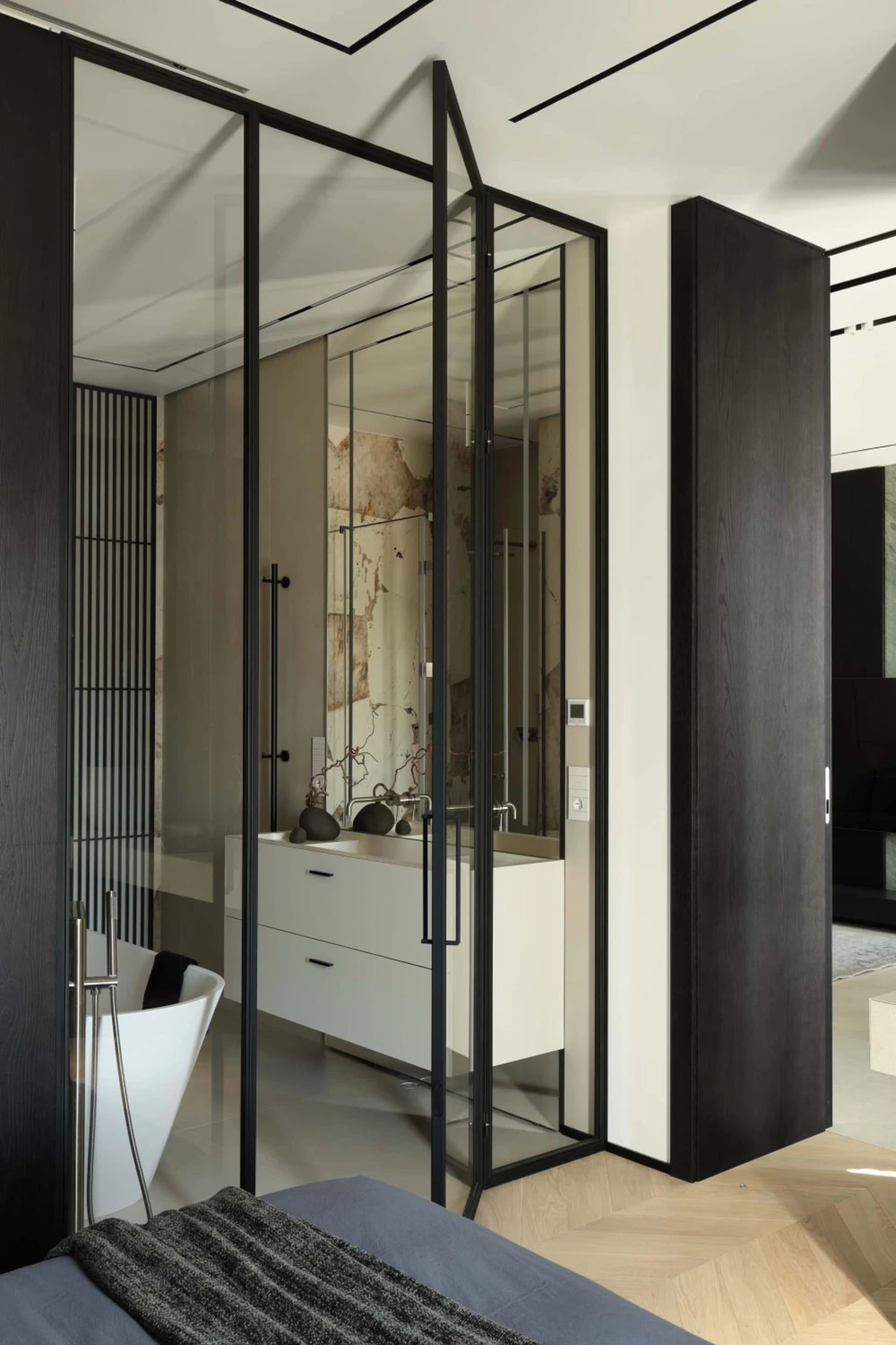 Modern ensuite bathroom viewed from bedroom, featuring black steel and glass folding partition, white floating vanity, marble accent wall, and freestanding white bathtub.