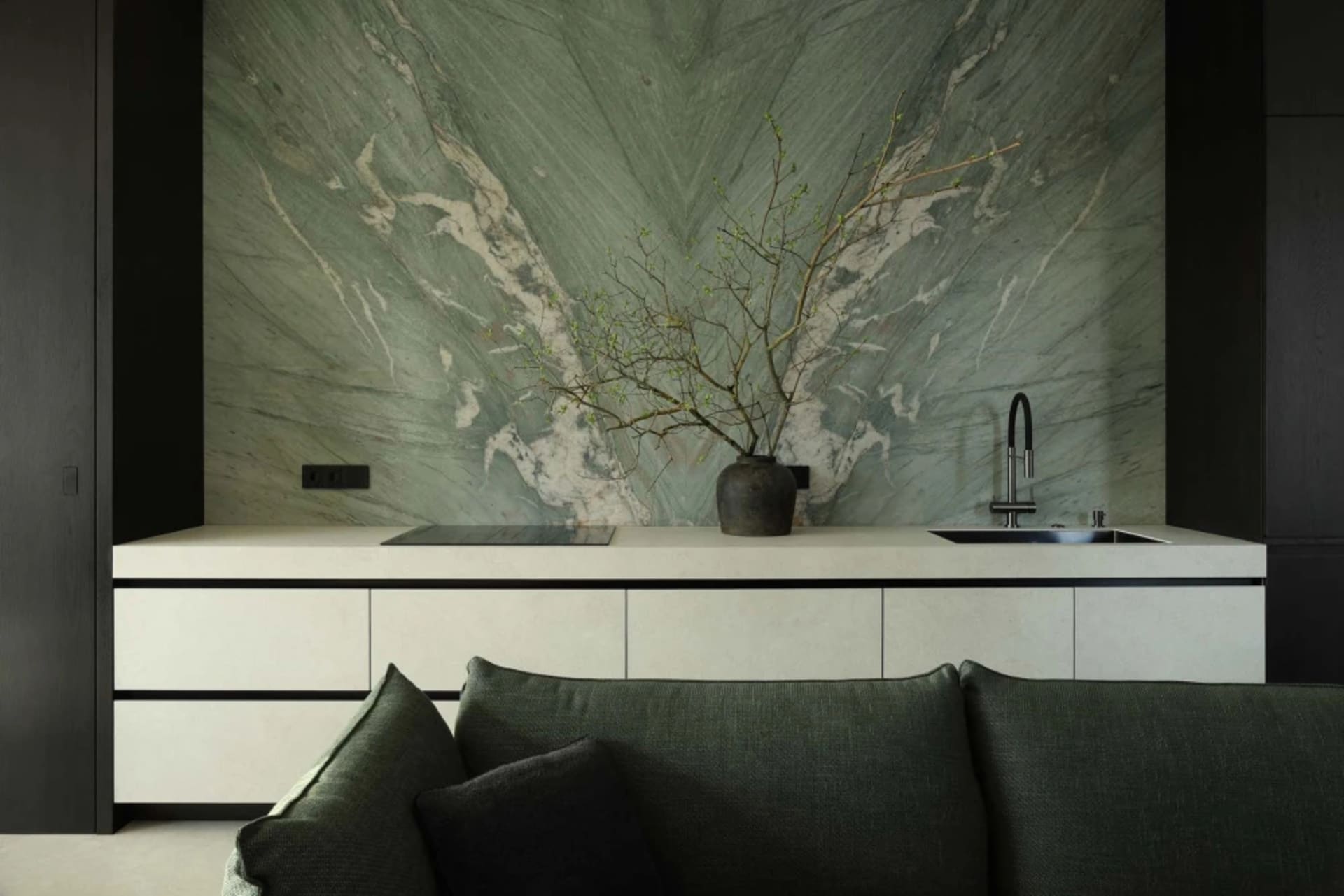 Modern open-plan kitchen and living area featuring a large sage green marble feature wall, white minimalist cabinetry, black fixtures, and a dark green sofa in the foreground.