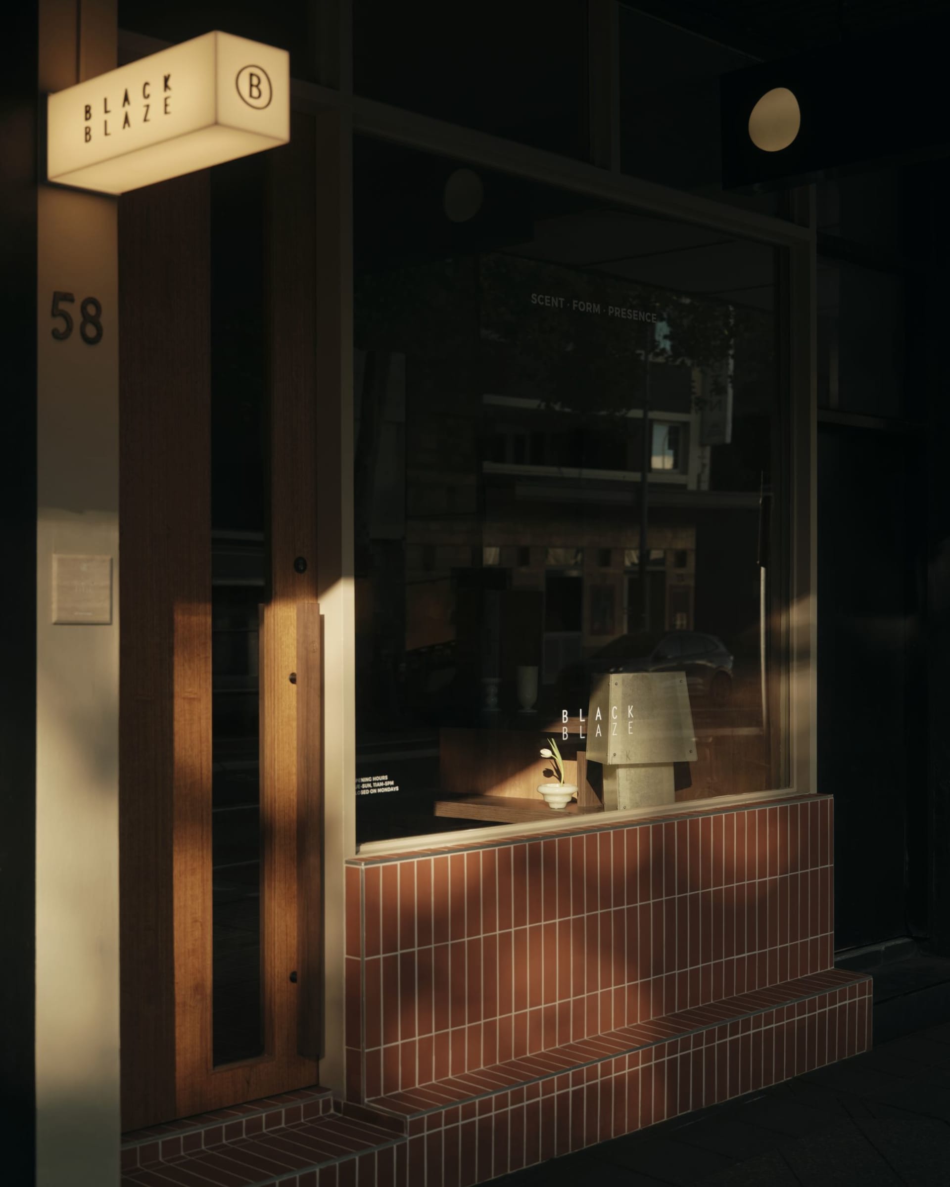 Night view of a modern storefront featuring a warm wooden door, terracotta tile wainscoting, illuminated Black Blaze sign, and a small floral arrangement in the window.