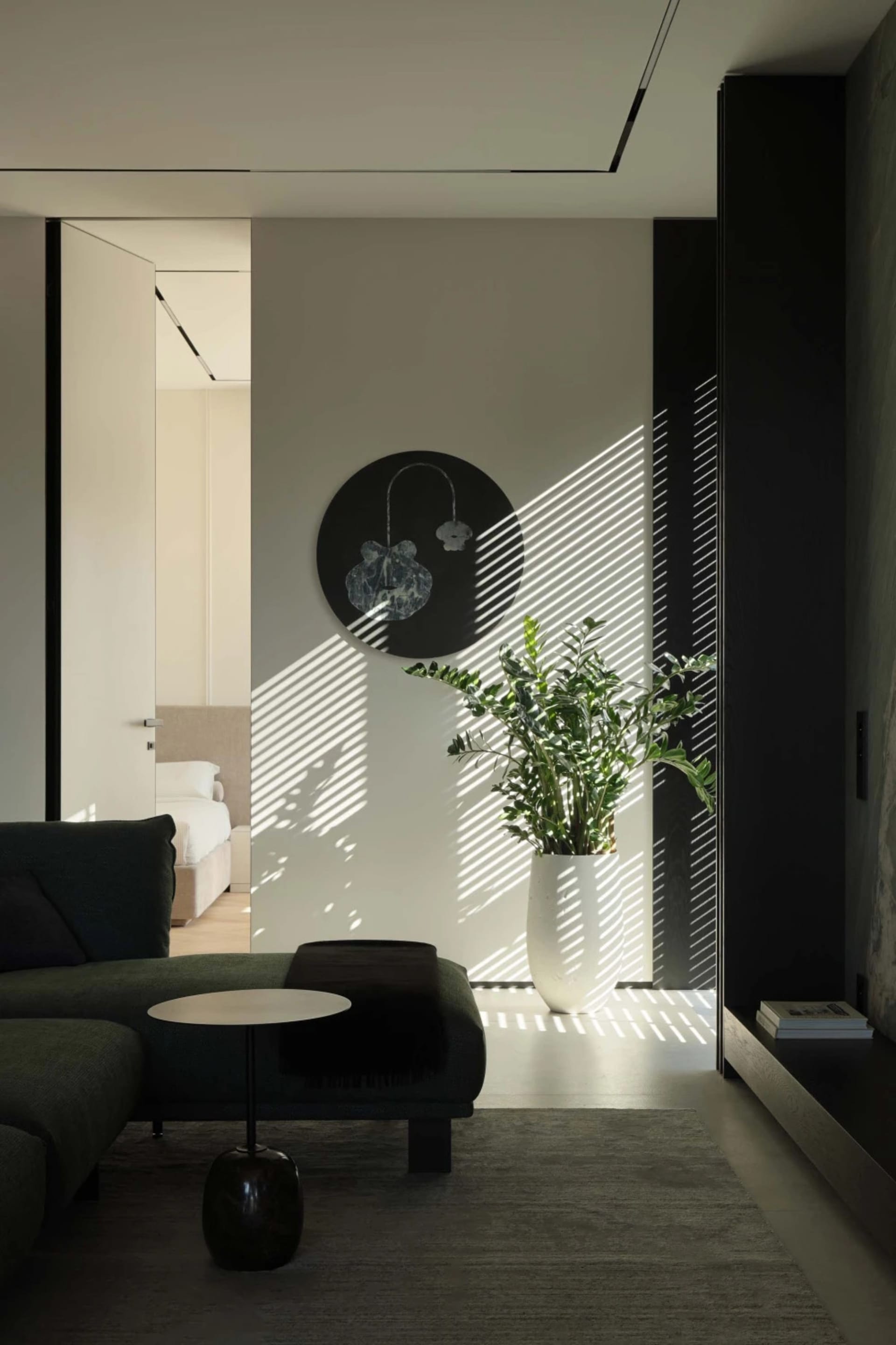 Modern minimalist living room featuring a dark green sectional sofa, round side table, large potted ZZ plant, circular wall art, and dramatic diagonal sunlight stripes on a light concrete wall.