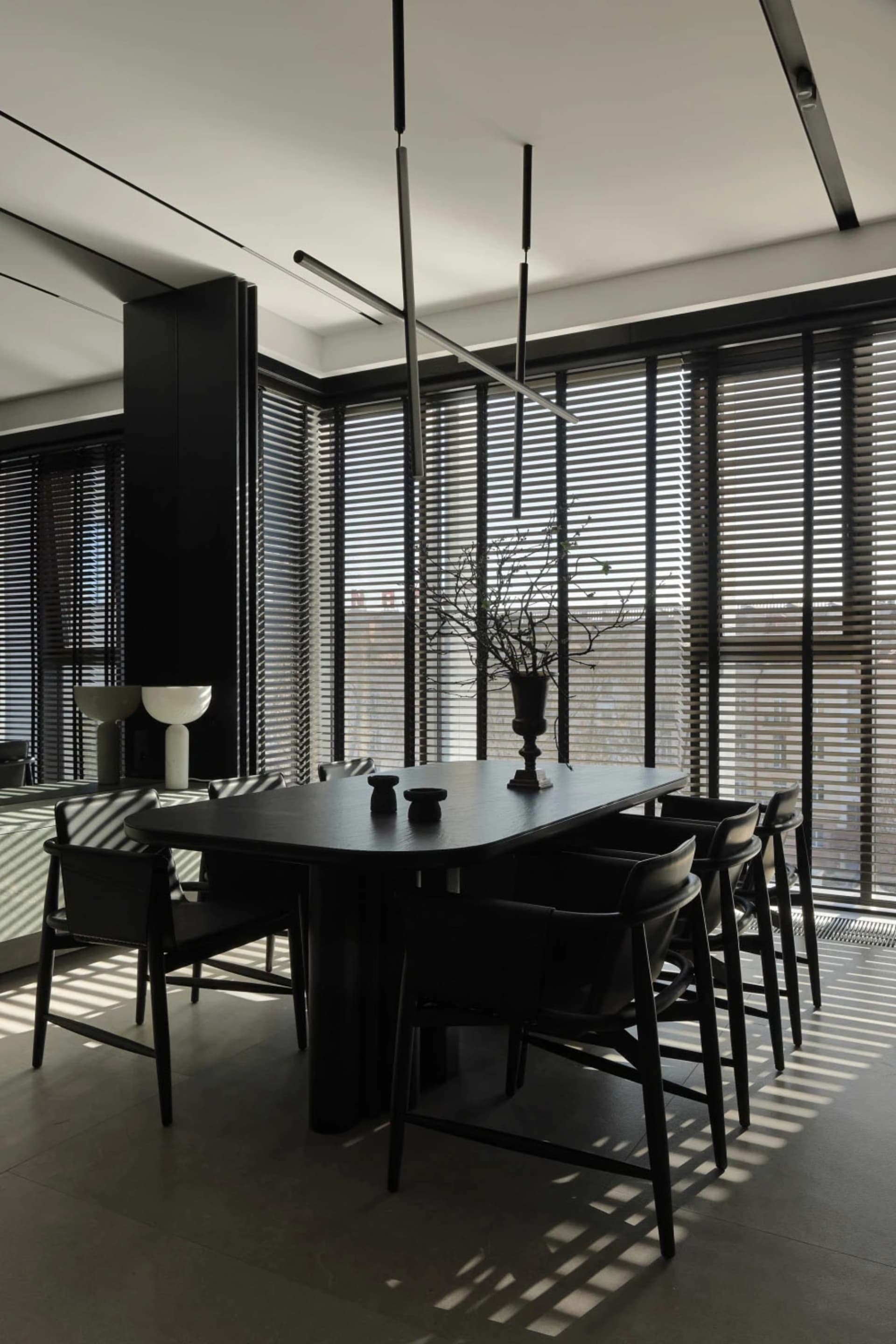 Modern minimalist dining room featuring a black oak rectangular table surrounded by six black leather chairs, dramatic linear pendant lights, large windows with horizontal blinds, and a tall black storage unit.
