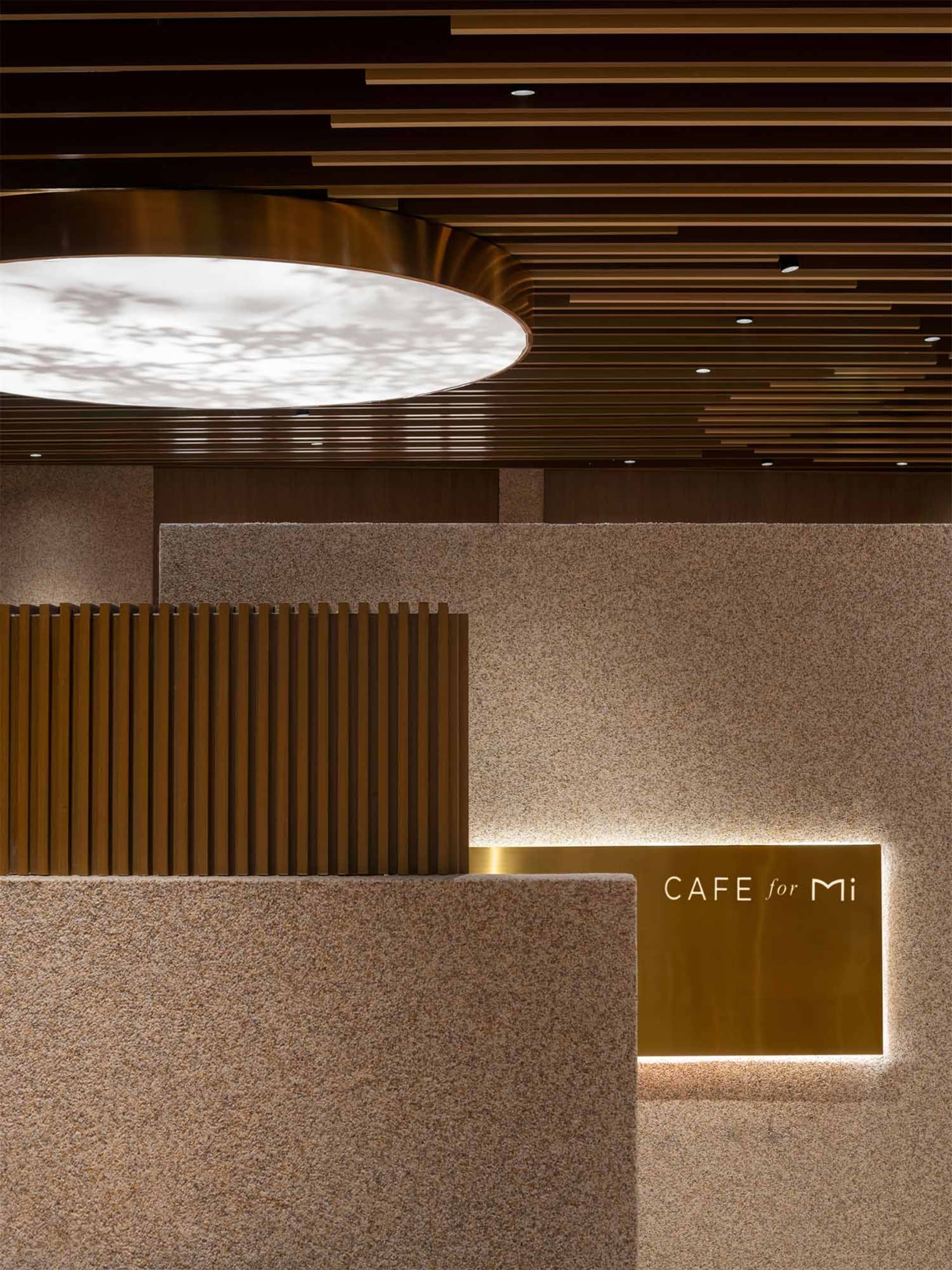 Modern cafe counter featuring speckled terrazzo walls, vertical wood slats, a large circular backlit ceiling fixture, and an illuminated brass sign reading 'CAFE for Mi'.