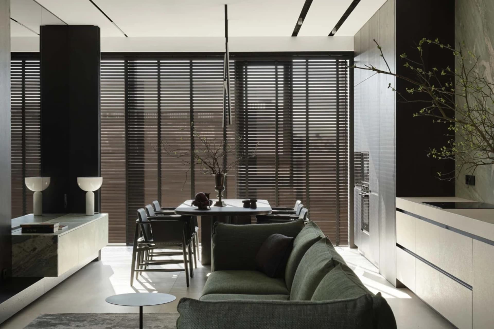 Modern minimalist open-plan dining and living room with sage green sofa, white marble dining table, dark wooden blinds, and integrated cabinetry bathed in natural sunlight.