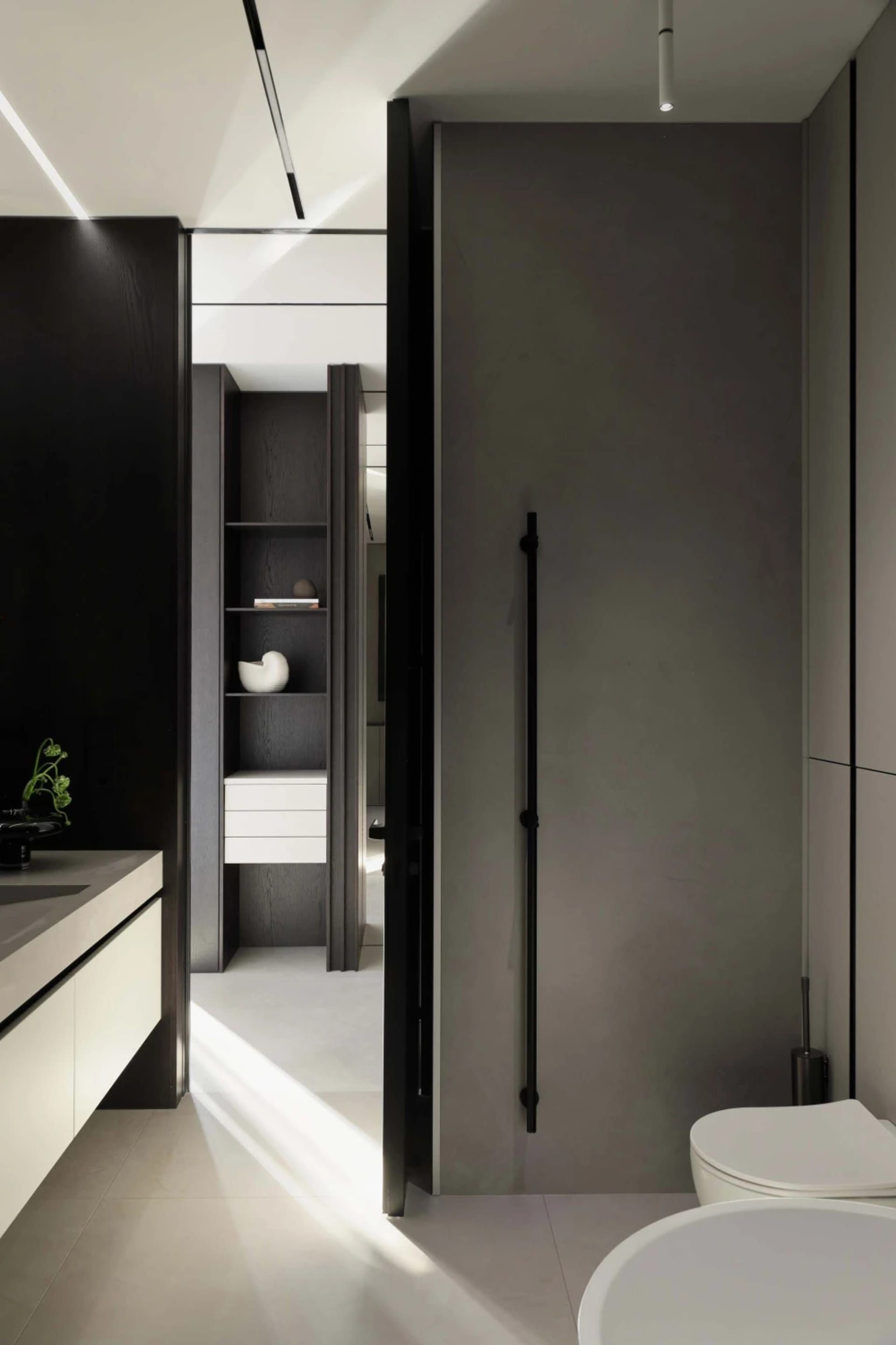 Modern minimalist bathroom interior with dark wood cabinetry, white floating vanity, built-in open shelving, matte black door, and white ceramic toilet under dramatic natural light.