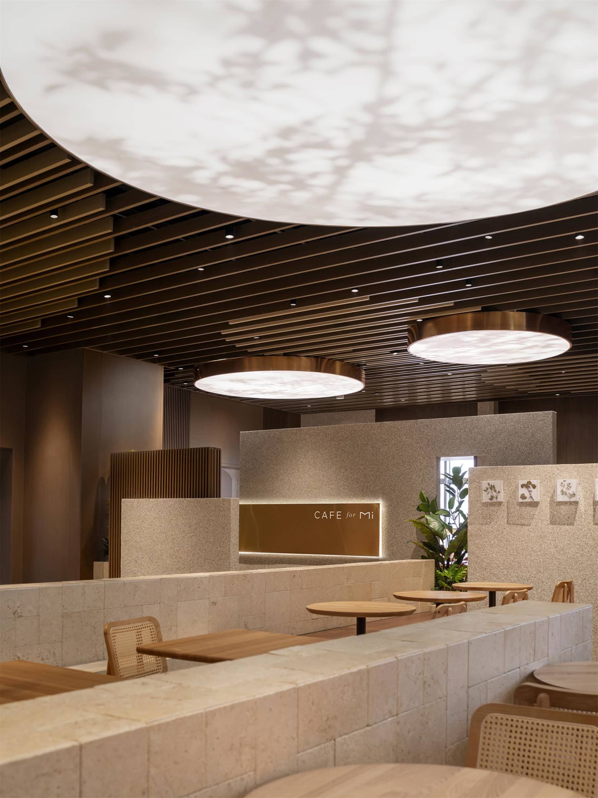 Modern café interior featuring light-colored terrazzo counters, wooden tables, rattan chairs, and large circular pendant lights with leaf-patterned projections on the curved wooden slat ceiling.