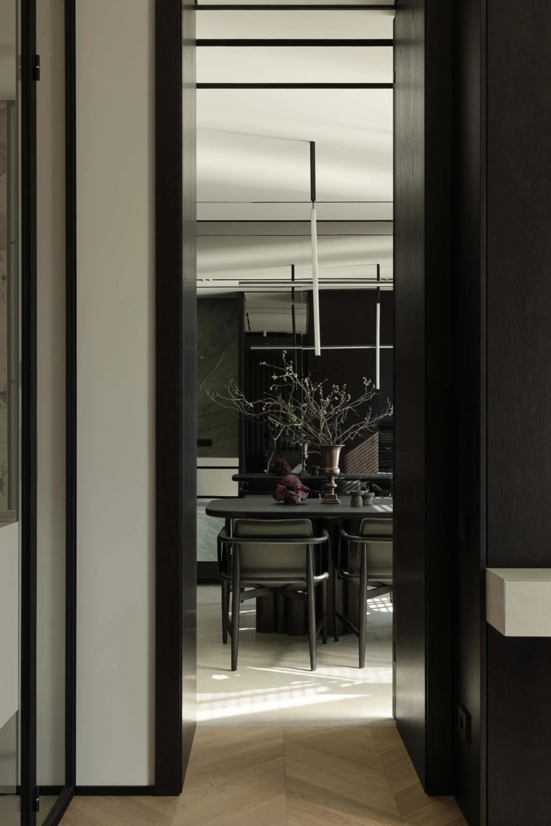 View from a hallway through a dark wood door frame into a modern minimalist dining room with dark table, leather chairs, pendant lights, and a large sculptural branch arrangement in a vase.