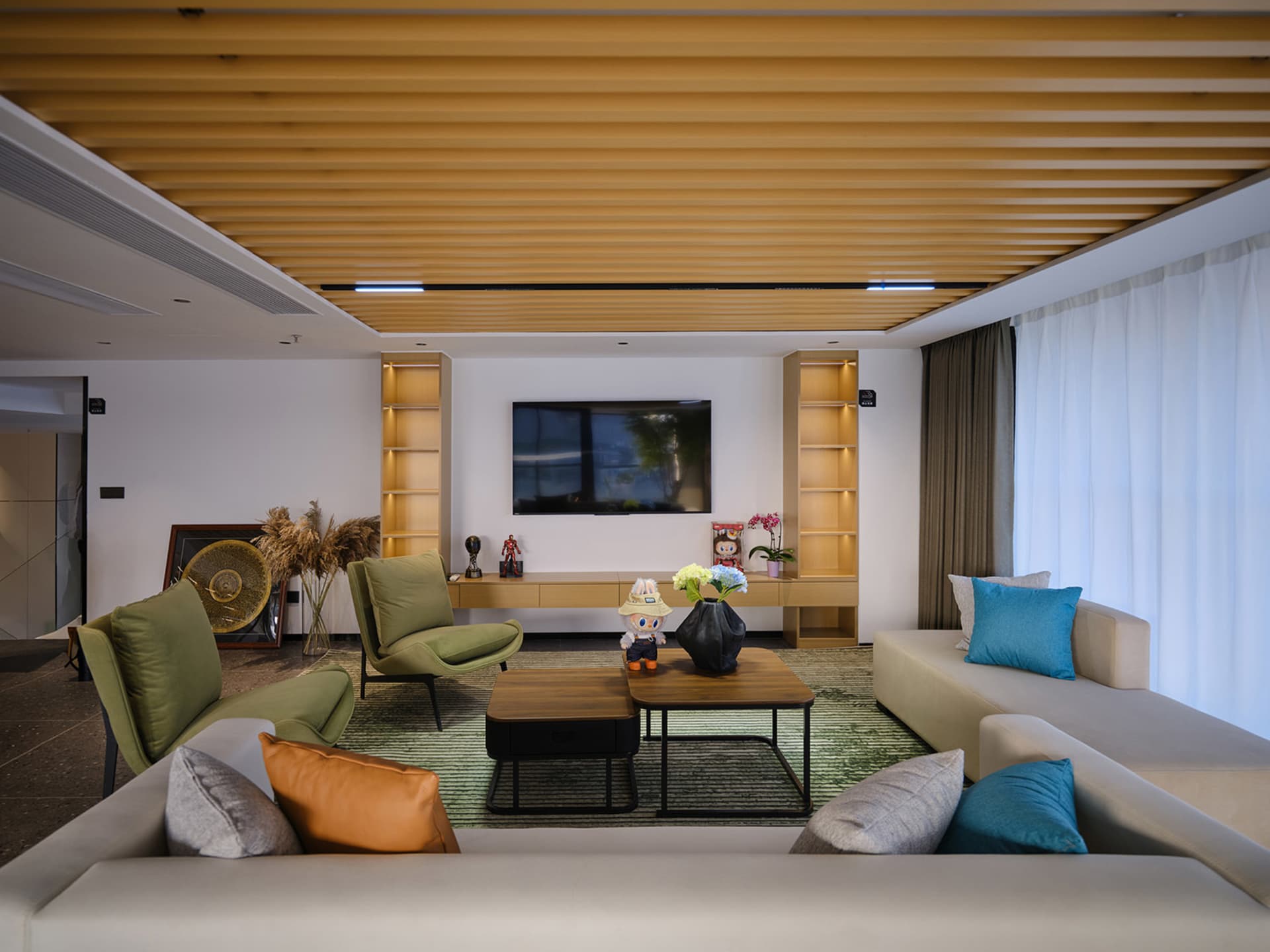 Modern living room with light wood slat ceiling, green velvet accent chairs, beige sectional sofa, wooden coffee tables on a green striped rug, and built-in illuminated shelving units.