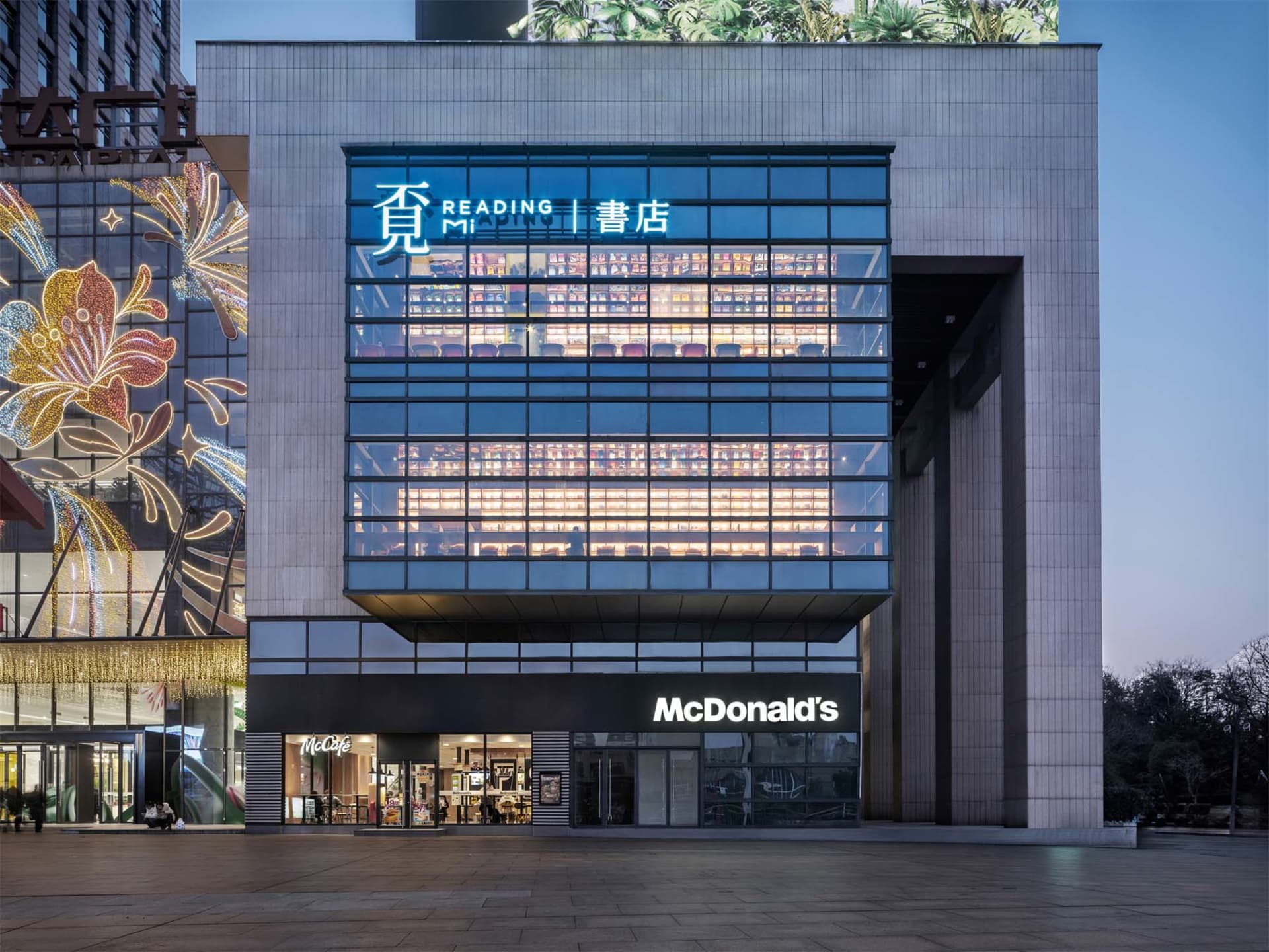 夜間拍攝的現代商業建築，特色為發光雙層挑高玻璃閱讀室，內有滿牆書架與甕閱讀書店招牌，下方為麥當勞餐廳，旁邊建築有節慶花朵燈飾。