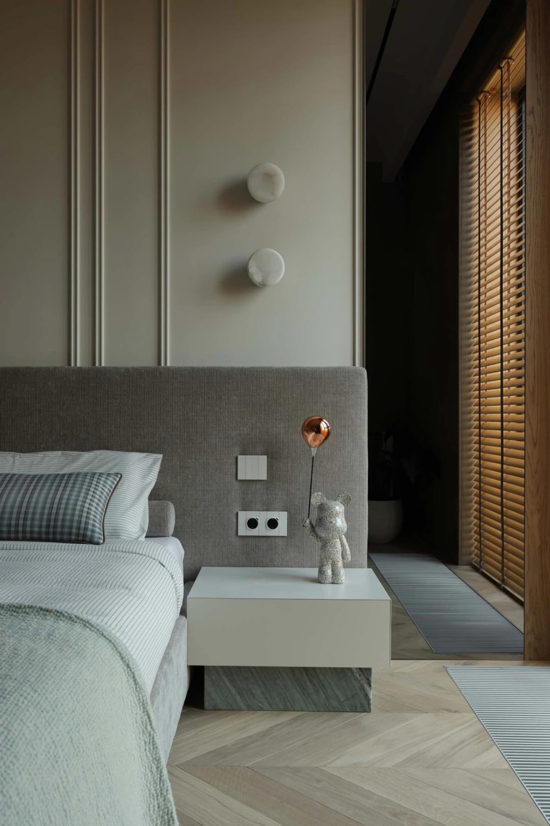 Modern minimalist bedroom featuring a grey upholstered headboard, white and marble bedside table with a copper balloon dog lamp, herringbone wood flooring, and wooden window blinds.