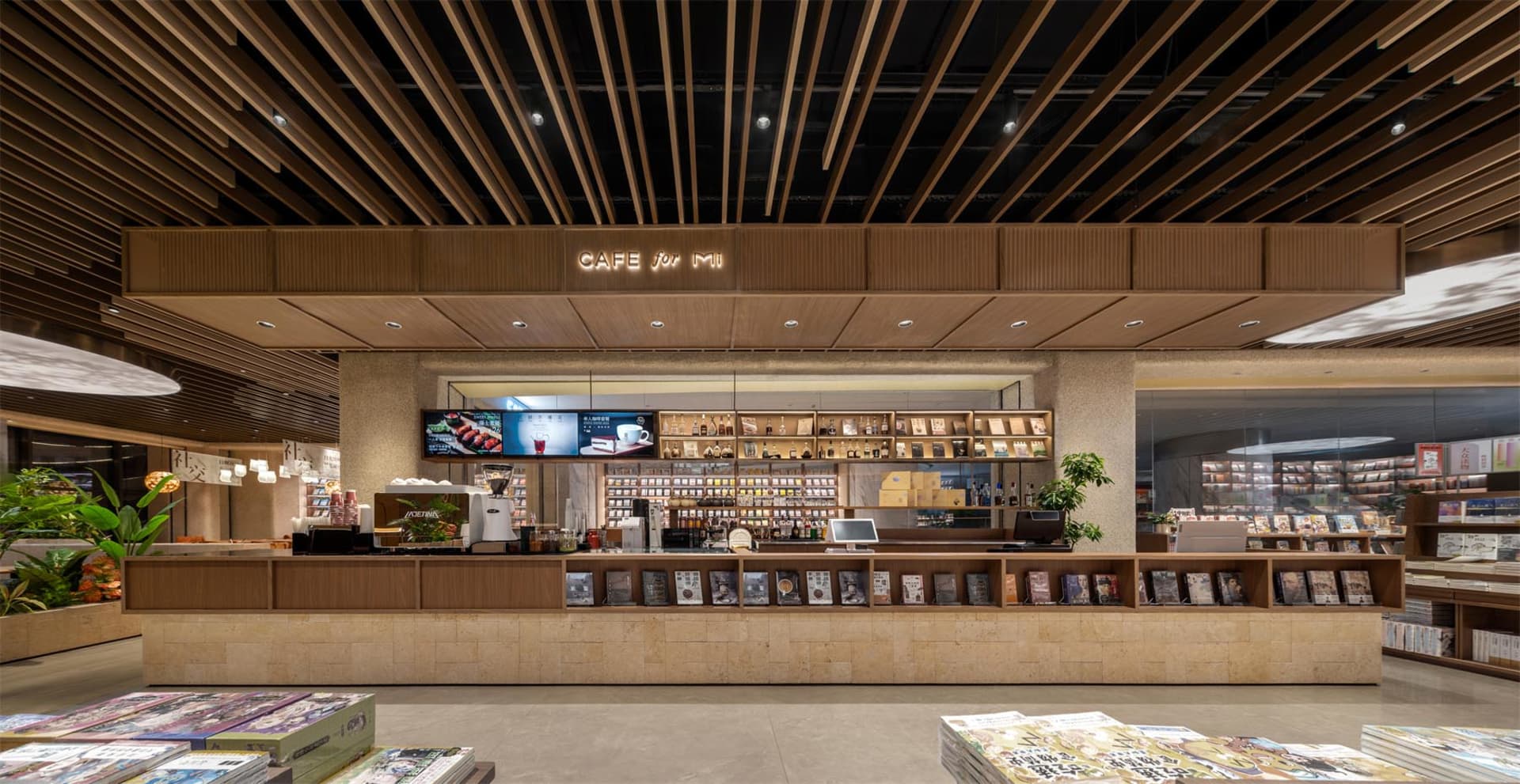 Modern bookstore cafe interior featuring a long travertine and wood service counter, linear wood slat ceiling, warm neutral palette, and integrated book displays.